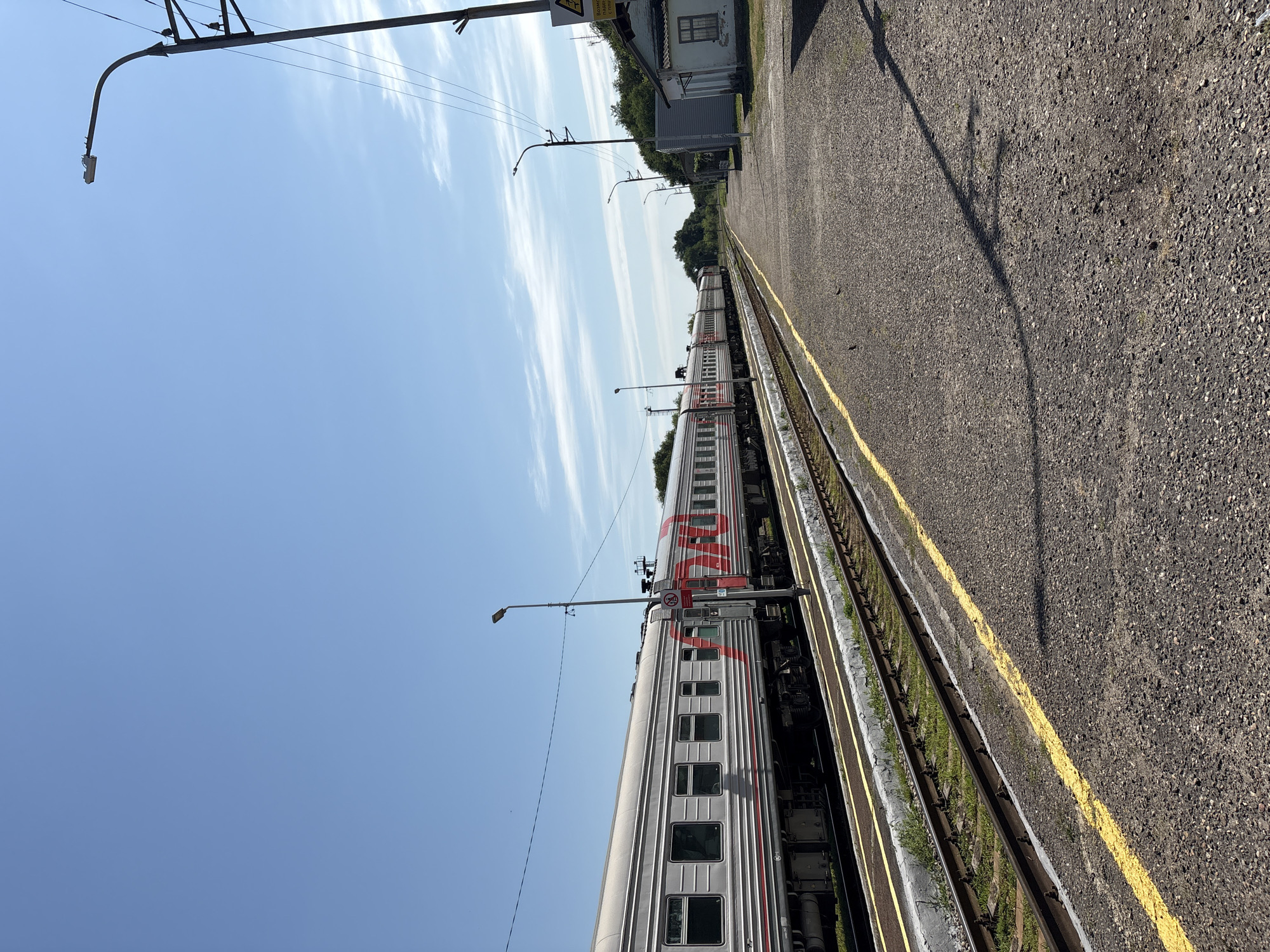 The image depicts a train at a small train station in the city of Izobilnyy, Russia. The train, composed of multiple silver and red carriages with Russian Railways (RZD) branding, is stopped on tracks to the left of the frame. The station platform, made of gravel with a prominent yellow line marking its edge, runs along the right side of the tracks. A single-story, light-colored building, likely a station house or shelter, is visible on the right side of the platform, with a window and a sloped roof. Several lampposts, including one with a curved arm, are present along the platform and in the background. The sky is clear blue with scattered white clouds. The environment appears to be rural or semi-rural, with trees and vegetation visible in the distance beyond the station building. The lighting suggests it is daytime, likely mid-morning or afternoon, given the clear sky and shadows. There are no people visible in the image.