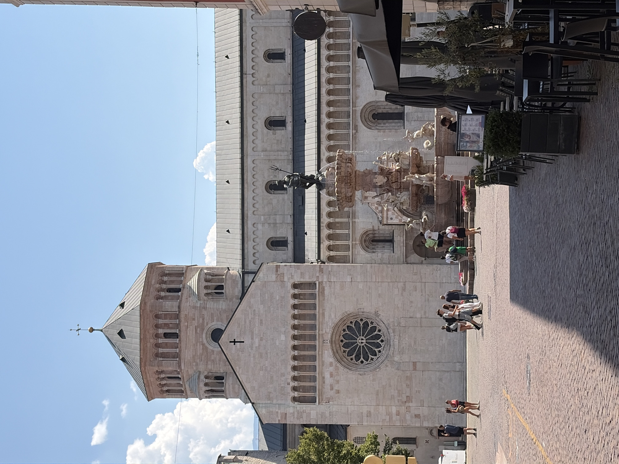 The image depicts a sunny day in Piazza Duomo in Trento, Italy, featuring the Trento Cathedral (Cattedrale di San Vigilio) and the Fountain of Neptune. The cathedral, a Romanesque-style building, dominates the background with its stone facade, octagonal tower, and rose window. The Fountain of Neptune is situated in the foreground, with its statue of the sea god holding a trident and surrounded by ornate sculptures.
Several people are visible in the square. A group of individuals are walking across the cobblestone pavement in the middle ground, some interacting with each other. On the right side of the image, outdoor seating for a cafe or restaurant is set up, with tables and chairs arranged under umbrellas.
The environment is a public square with a clear blue sky and scattered white clouds. The cobblestone pavement is a prominent feature of the ground. The overall scene conveys a lively atmosphere in a historic city center.