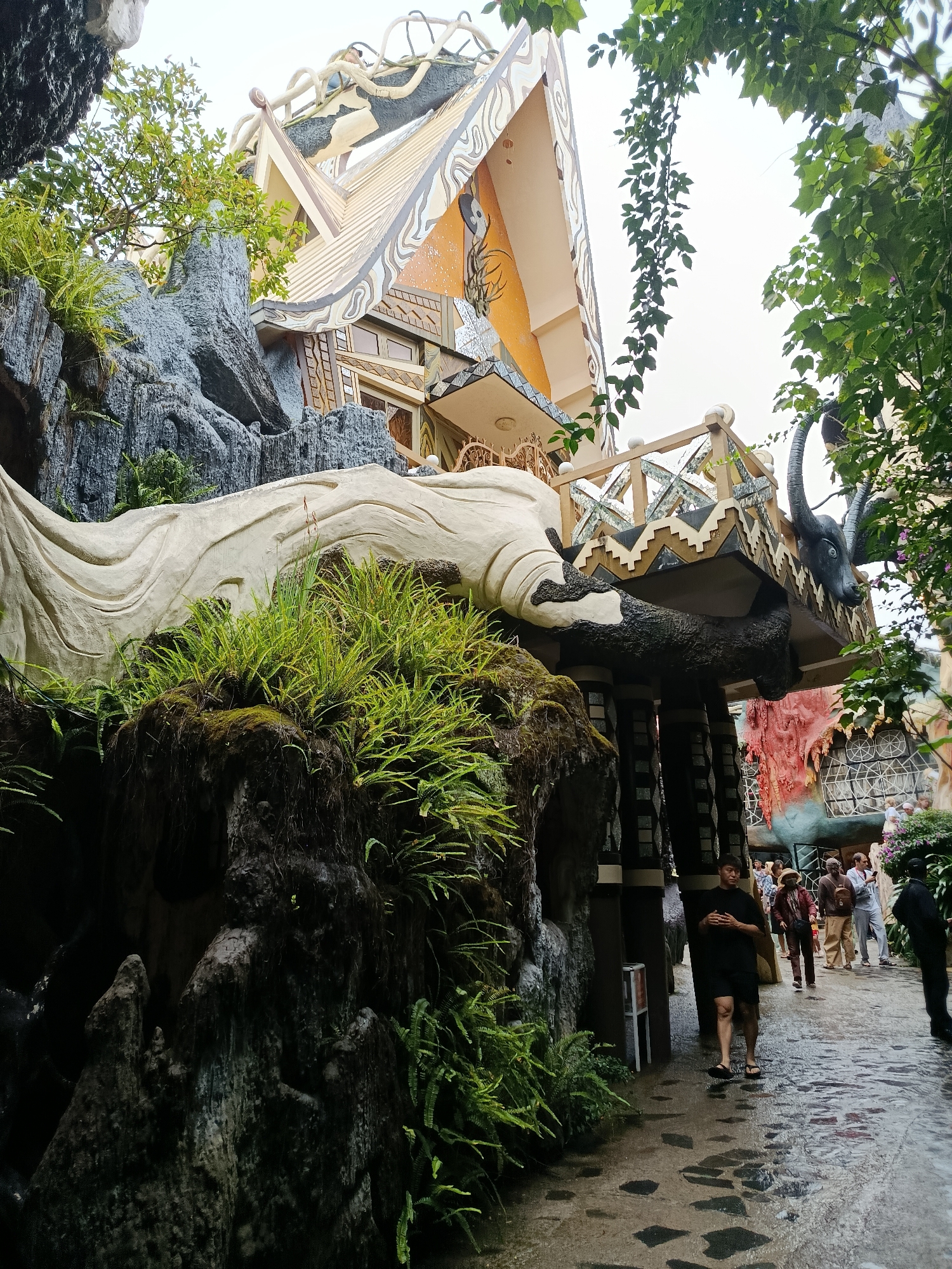 The image depicts the exterior of a unique, whimsical building with an unconventional architectural design. The building features a multi-layered roof with a distinctive curved shape, adorned with intricate patterns and textures. The facade incorporates elements resembling natural rock formations and sculpted figures, including a large, pale, wave-like structure with lush green ferns growing on it. A prominent dark sculpture of an animal head with large horns protrudes from an elevated section of the building. The pathway leading to the structure is paved with irregular stones and is wet, suggesting recent rain. Several people are visible on the pathway, walking towards or standing near the building, dressed in casual attire. Green foliage, including vines and trees, frames the scene, contributing to an overall surreal and artistic ambiance. The lighting is overcast, indicating a cloudy day.