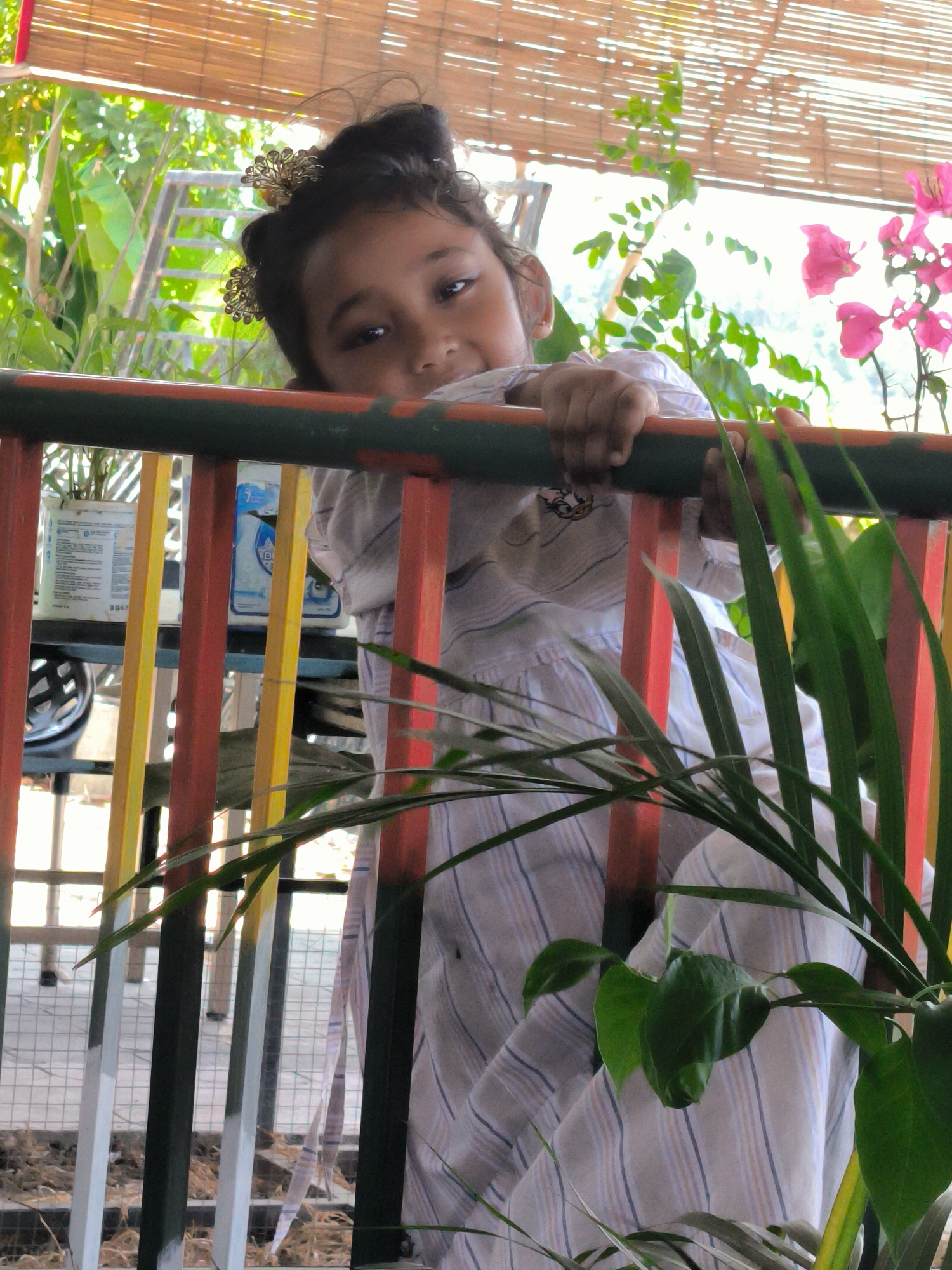 A young child with a hair accessory is leaning over a brightly painted railing in what appears to be a garden or patio. The location is Matang, Indonesia. The child wears a striped shirt. Plants and flowers are visible in the background. Various objects such as a bottle and a helmet are on a table behind the railing. The railings are painted in green, red, yellow and white. A screen is at the top of the image.