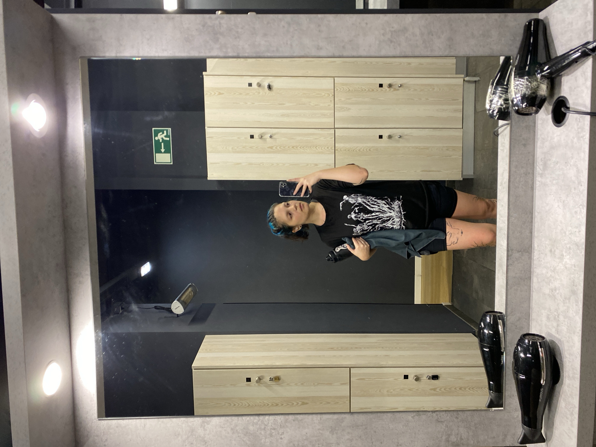 This vertical image shows a young woman taking a selfie in a large mirror within what appears to be a changing room or locker room. The woman, reflected in the mirror, has blue-dyed hair and a neutral expression. She is wearing a black t-shirt with a white graphic design and black shorts, with tattoos visible on her legs. She holds a smartphone in her right hand, capturing her reflection, and a black water bottle along with a dark cloth or towel in her left hand.
The setting features textured gray walls and a dark, possibly black, background visible beyond the mirror's reflection. Prominently reflected in the mirror are light-colored wooden lockers or cabinets with silver handles and small black square locks, indicating a storage area. On the right side, two black wall-mounted hair dryers are visible, with the brand name "FURIOUS" clearly legible on one. Bright, circular wall-mounted lights illuminate the space. Further details reflected in the mirror include a green emergency exit sign depicting a figure running down a ramp with an arrow, and a white cylindrical object, possibly a portable heater or diffuser, on a dark surface in the background.