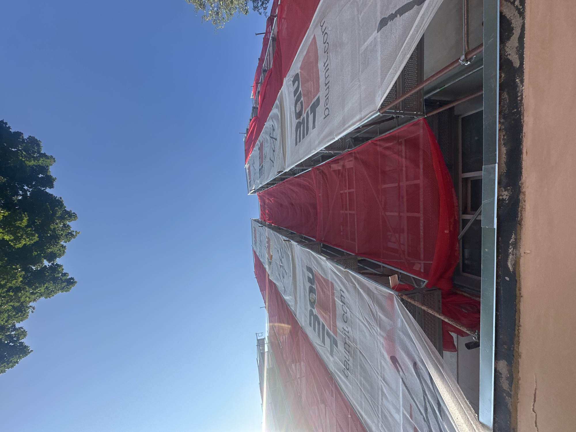 The image shows a close-up, low-angle view of a building under renovation, covered in red and white tarps. The tarps are stretched over scaffolding that encloses the upper floors of the structure. The white tarps feature large, black and red logos that appear to be for a company named "BAUMIT" with the website "baumit.com" printed below it. The red tarps are a solid color and are visible beneath the white ones. The sky above is clear and blue, with the tops of green trees visible in the upper left and right corners of the frame. The bottom of the image shows the edge of the building's roof or upper facade, with a strip of metal trim. The sun is shining brightly, creating a lens flare effect on the left side of the image.