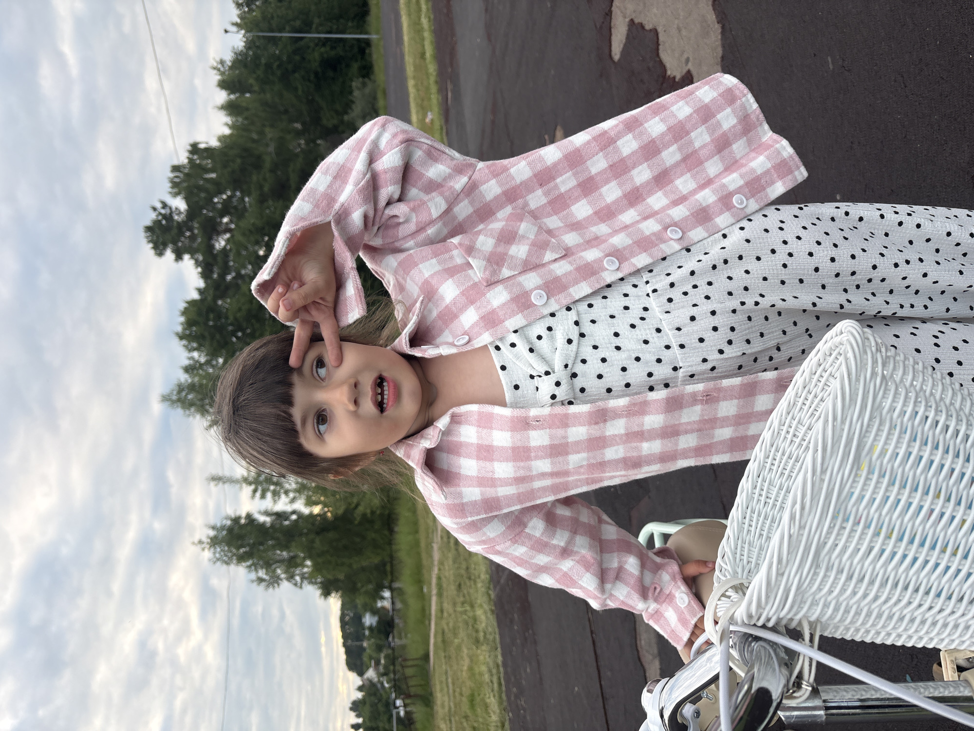 A young girl with dark hair and brown eyes is positioned in the center of the frame, her body angled towards the viewer. She is wearing a white and pink gingham-patterned shirt over a white dress with black polka dots. The girl is making a peace sign with her right hand, held up near her eye. Her mouth is slightly open, revealing her teeth. She appears to be sitting on a bicycle, with a white wicker basket visible in the lower right portion of the image. The background consists of a paved surface, green grass, and trees. The sky is cloudy, suggesting it might be overcast or late in the day. The overall emotion conveyed is playful and cheerful. There is no visible text in the image. The location context is Chaikovsky, Russia.