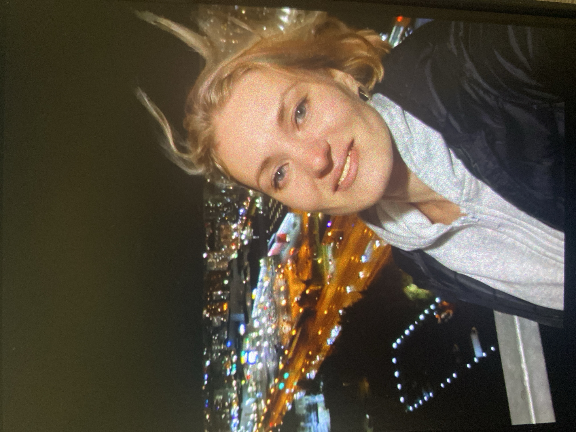 A young woman is captured in a selfie-style photograph at night, taken from a high vantage point overlooking a sprawling city. She is positioned with her head tilted, smiling directly at the camera, conveying a cheerful and engaging expression. Her blonde hair appears windswept or dynamically styled, framing her face. She has light-colored eyes and is wearing a dark jacket over a light grey hooded sweatshirt.
The background is dominated by a vibrant cityscape at night, with countless blurry, colorful lights creating a bokeh effect. These lights suggest illuminated buildings, streets, and vehicles, indicative of a large urban environment. A dark structure, possibly a railing or part of a building, is visible in the bottom right corner, suggesting she is on a balcony, rooftop, or observation deck. The scene is illuminated primarily by the city lights and what appears to be a light source or flash directed at the woman. The overall mood is bright and joyful against the dark, glittering backdrop of the city at night. The location is Saint Petersburg, Russia. There is no visible text in the image.