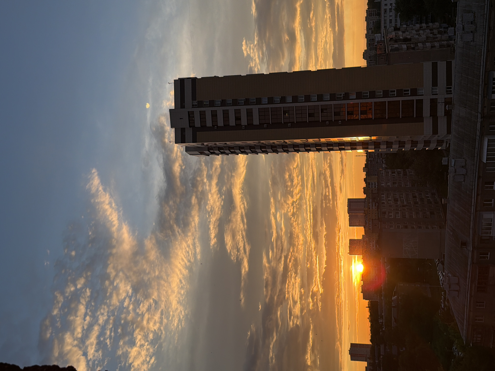 The image displays a vibrant sunset over an urban landscape. A tall, modern multi-story building, likely residential, stands prominently in the center-right foreground. Its light-colored facade features numerous windows, some reflecting the warm, golden light of the setting sun.
The sky is a dominant feature, filled with scattered, elongated clouds that are brilliantly illuminated by the low sun. These clouds exhibit a palette of orange, yellow, and soft gray hues, particularly dense in the middle and lower left sections of the frame. In the upper left quadrant, a faint, pale moon is visible against the lighter blue expanse of the sky.
The setting sun is visible as a bright, glowing orb on the horizon, positioned behind a dense cluster of city buildings that form a dark silhouette against its intense light. The cityscape extends into the distance, showing numerous other multi-story structures. The overall scene captures the dramatic transition from day to night in an urban environment. No visible text is present in the image.