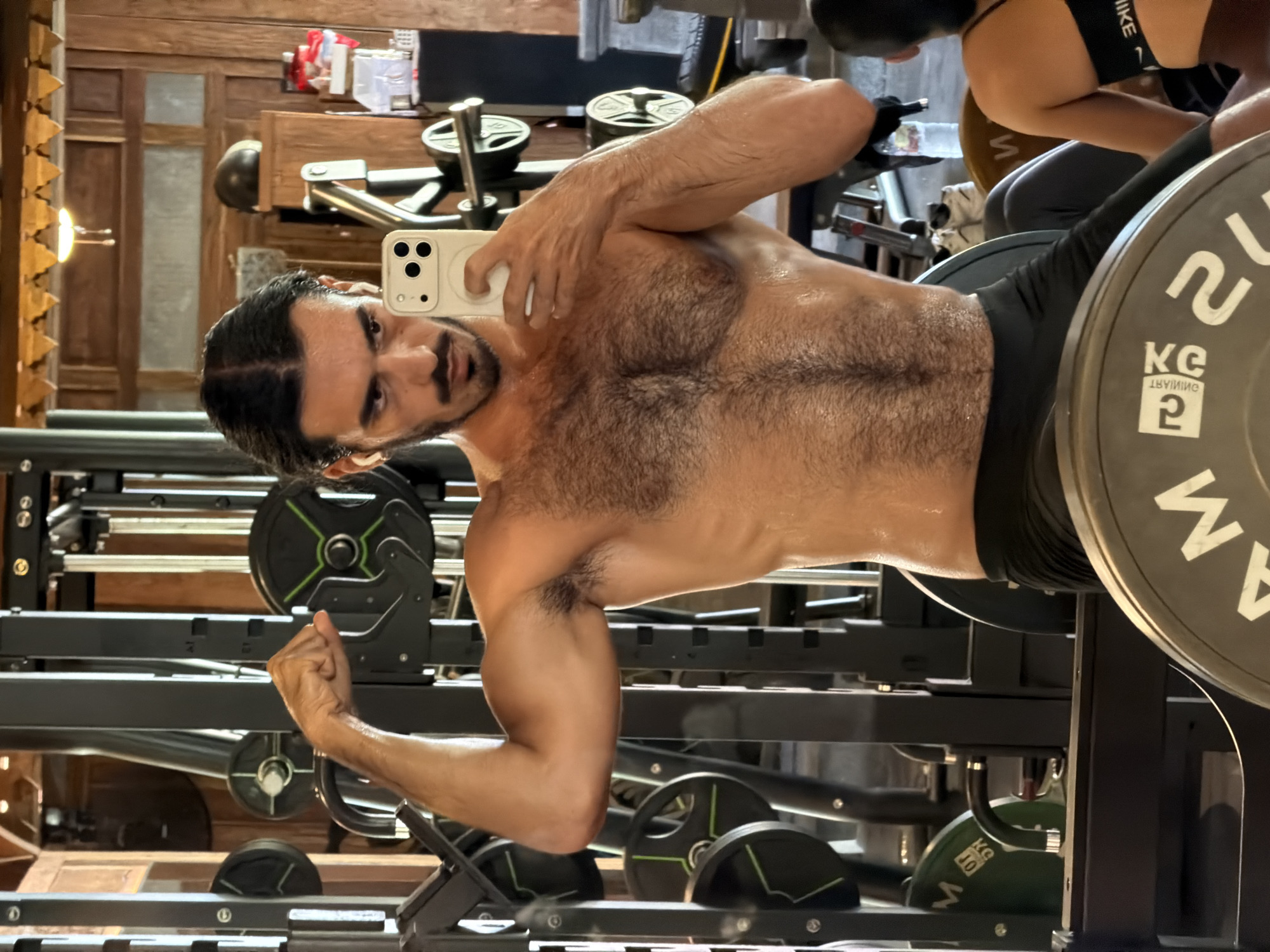 A shirtless man is taking a selfie in a gym in North Kuta, Indonesia. He is standing in front of a weight rack with plates on it. Another person wearing a Nike top is visible in the background.