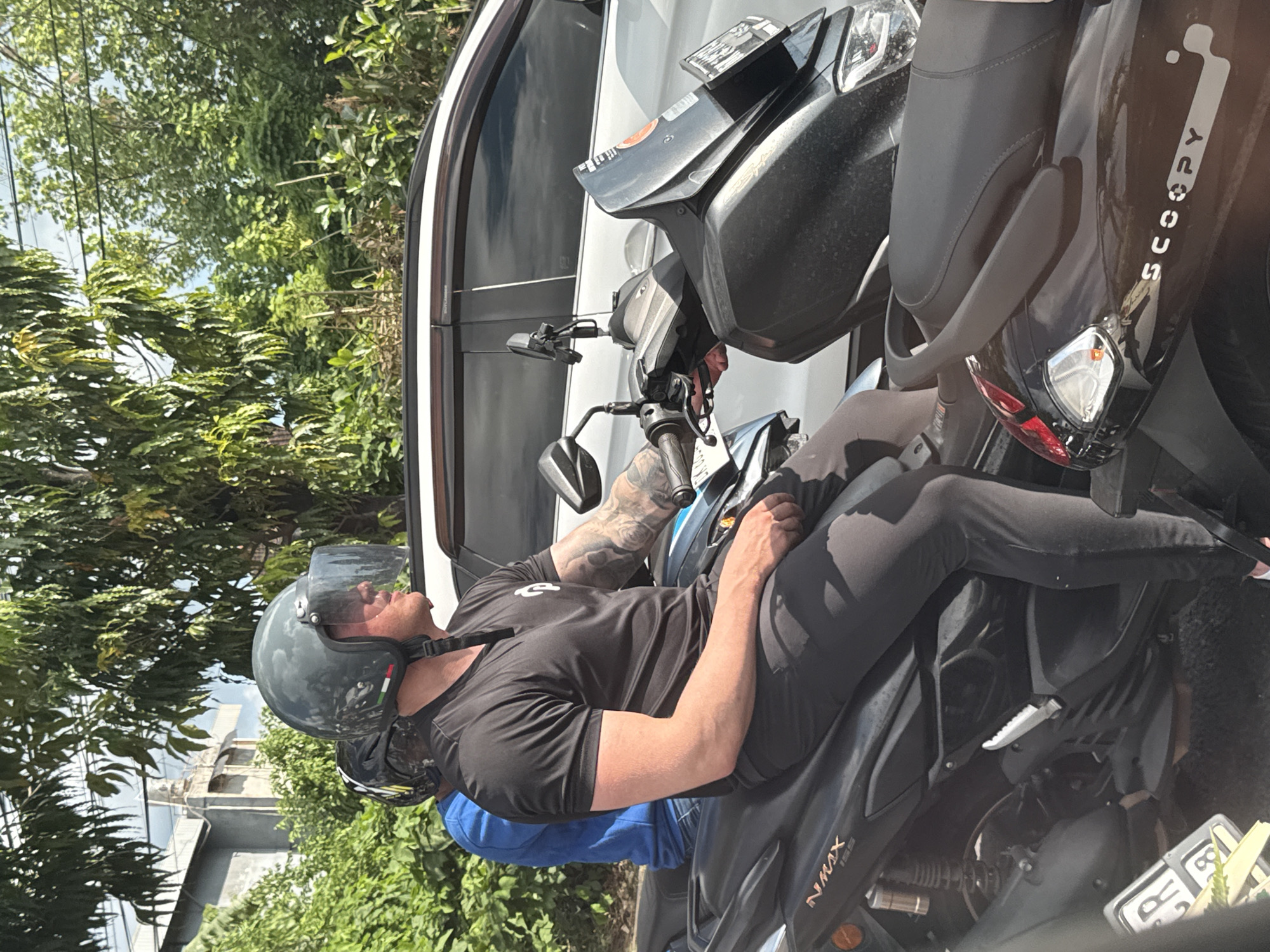 A medium shot features a person seated on a black NMAX motorcycle. The person, wearing a black t-shirt, black pants, and a helmet with a clear visor, has tattooed arms and is positioned between two parked motorcycles, one labeled "SCOOPY." The motorcycles are parked beside a white van. The setting appears to be a street scene in North Kuta, Indonesia, indicated by tropical vegetation in the background.