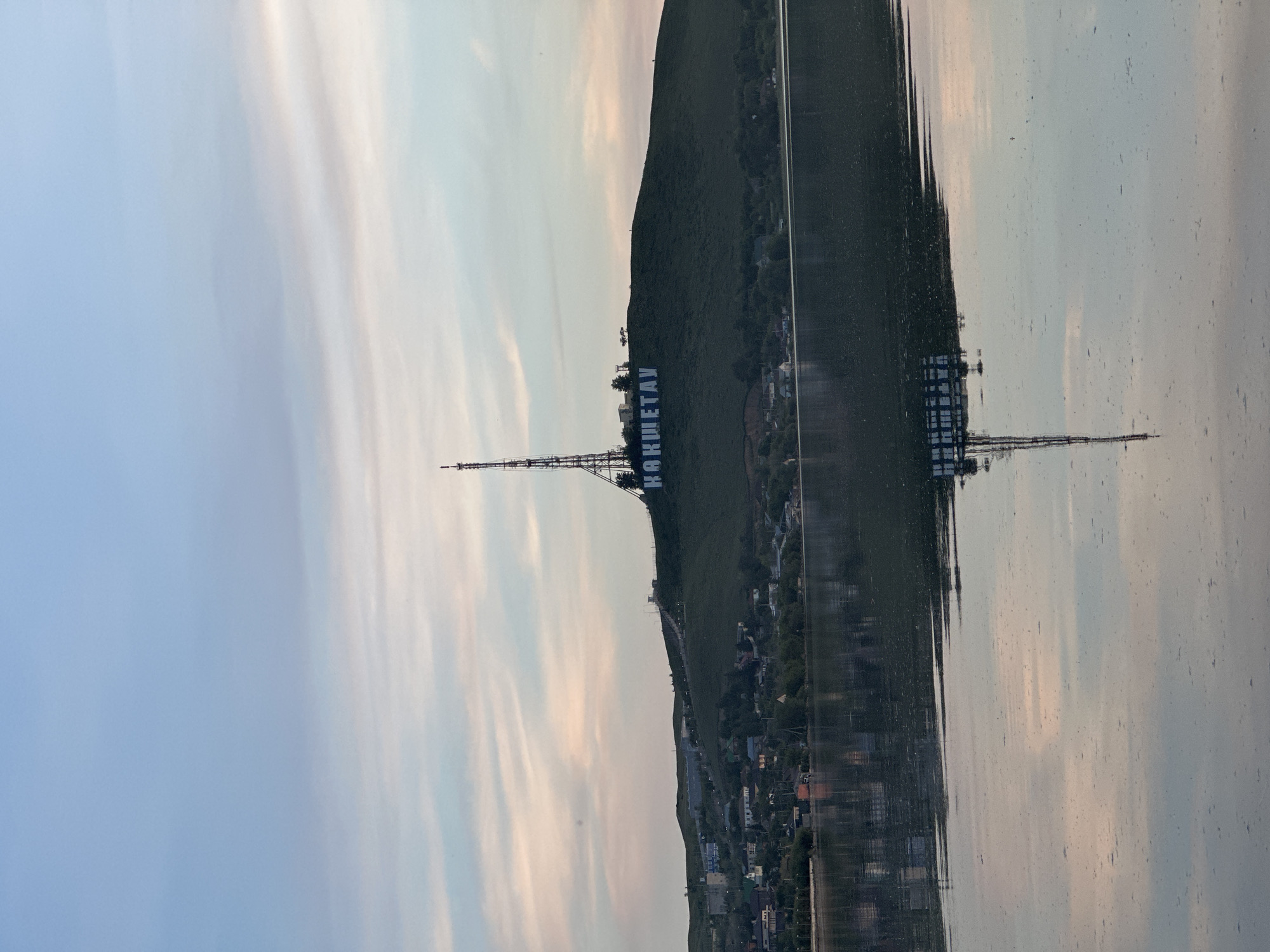 The image shows a landscape with a hill on which the word "KOKSHETAU" is written in large white letters in Cyrillic script. A tall communication tower is positioned nearby. A town or city is located at the base of the hill. The scene appears to be reflected in a body of water, indicating a lake or river in the foreground. The sky is slightly cloudy with light colors. The general location is the city of Kokshetau, Kazakhstan, evidenced by the lettering on the hill.