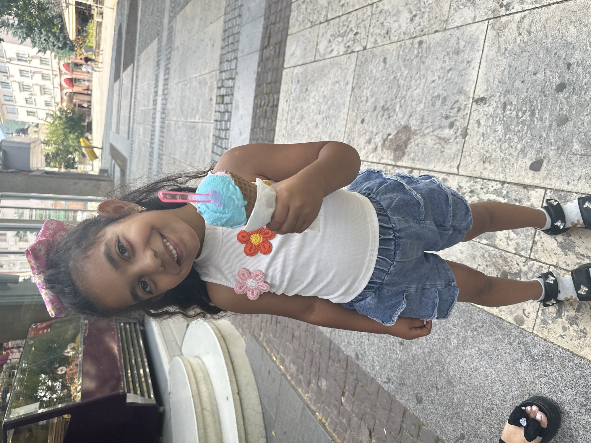 A young girl with dark, curly hair and a pink floral hair bow is smiling and holding a blue and brown ice cream cone. She is wearing a white tank top with two crocheted flowers, one orange and one pink, and a denim skirt. She has on white socks with a floral pattern and black sandals. The girl is outdoors, standing on a stone-tiled walkway. In the background, there are buildings and trees. The image appears to be taken during the day, as indicated by the bright lighting. The girl's expression suggests happiness and enjoyment, likely from eating her ice cream. There is no visible text in the image.