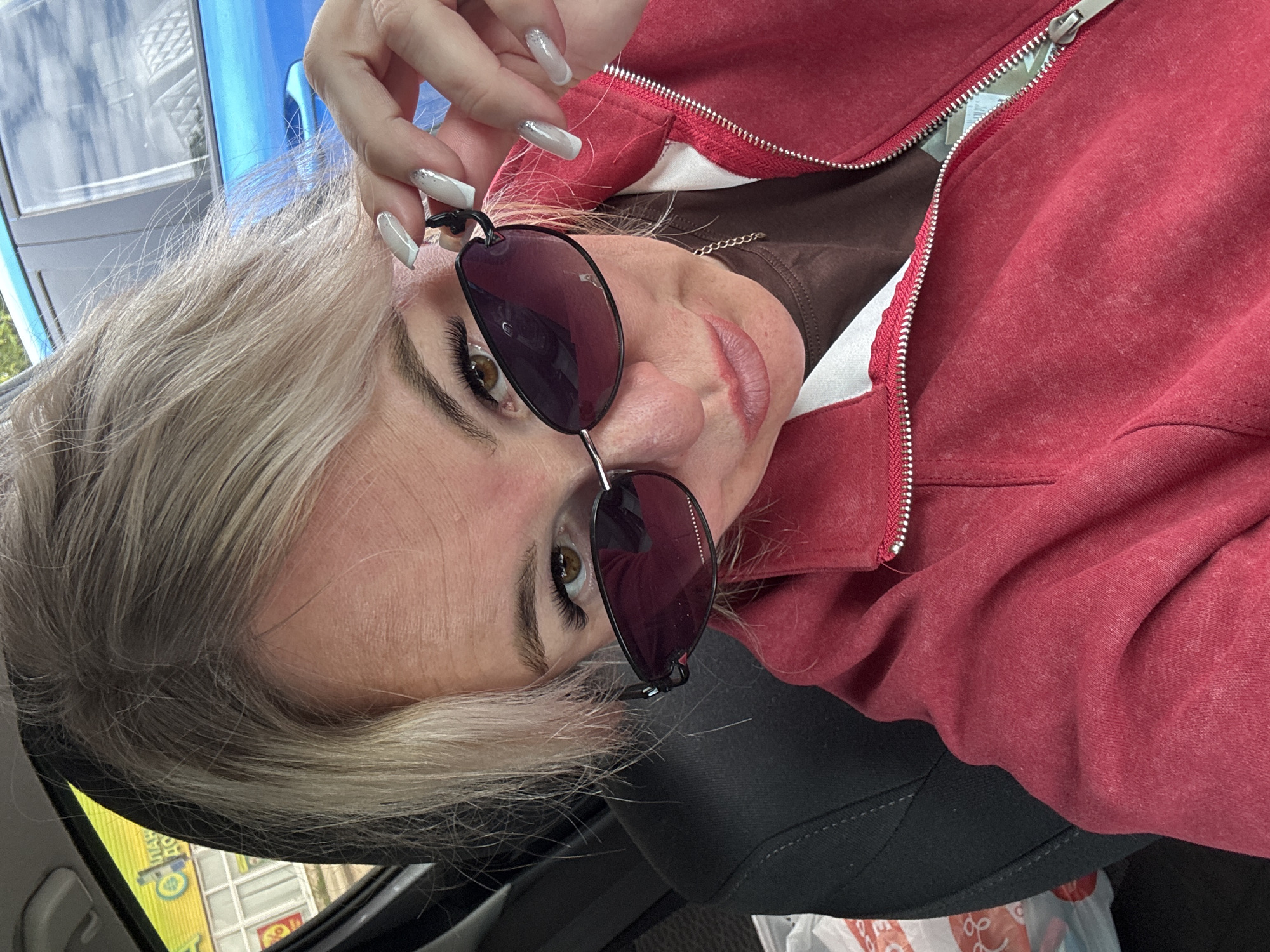 The image shows a close-up, self-taken portrait of a blonde woman with sunglasses, wearing a red zippered jacket over a brown shirt, inside a vehicle. The woman is holding her sunglasses with her right hand, revealing her eye and part of her face. Her nails are painted in a light color with silver glitter accents. The background shows the interior of a car, with a blue car visible through the window. The scene is likely in the city of Chastyye, Russia, as indicated by text on a yellow sign in the background which reads "ЧАСТЫЕ" (Chastyye).