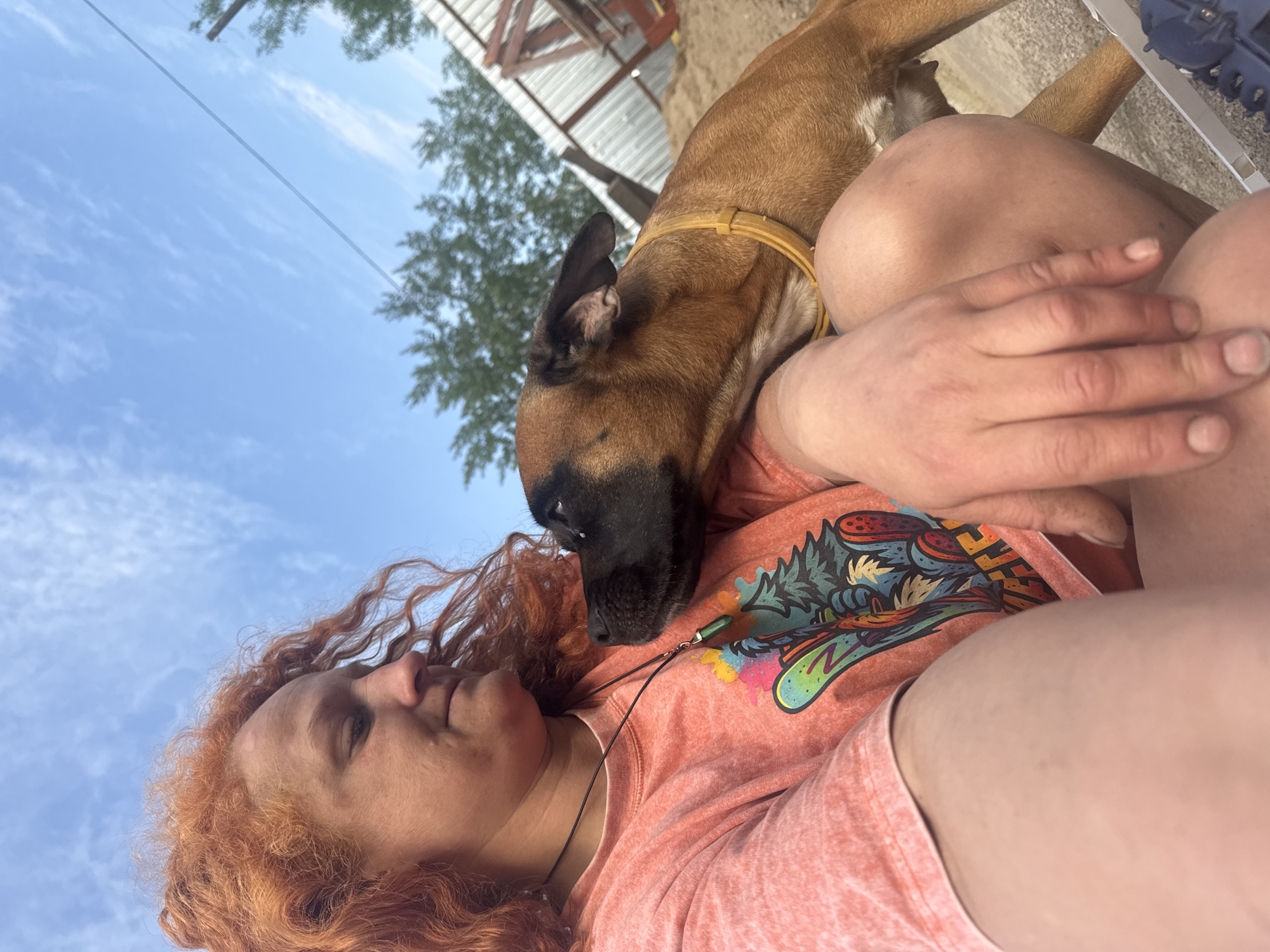 A woman with curly red hair lies reclined outdoors with a brown dog resting on her. The woman wears a coral-colored t-shirt with a colorful graphic. She is wearing a necklace with a small green stone pendant. Her eyes are closed, and her face is turned towards the sky. The dog has a black muzzle and is wearing a yellow collar. A building and trees are visible in the background. The sky is blue with sparse clouds. The image location is Chaikovsky, Russia.