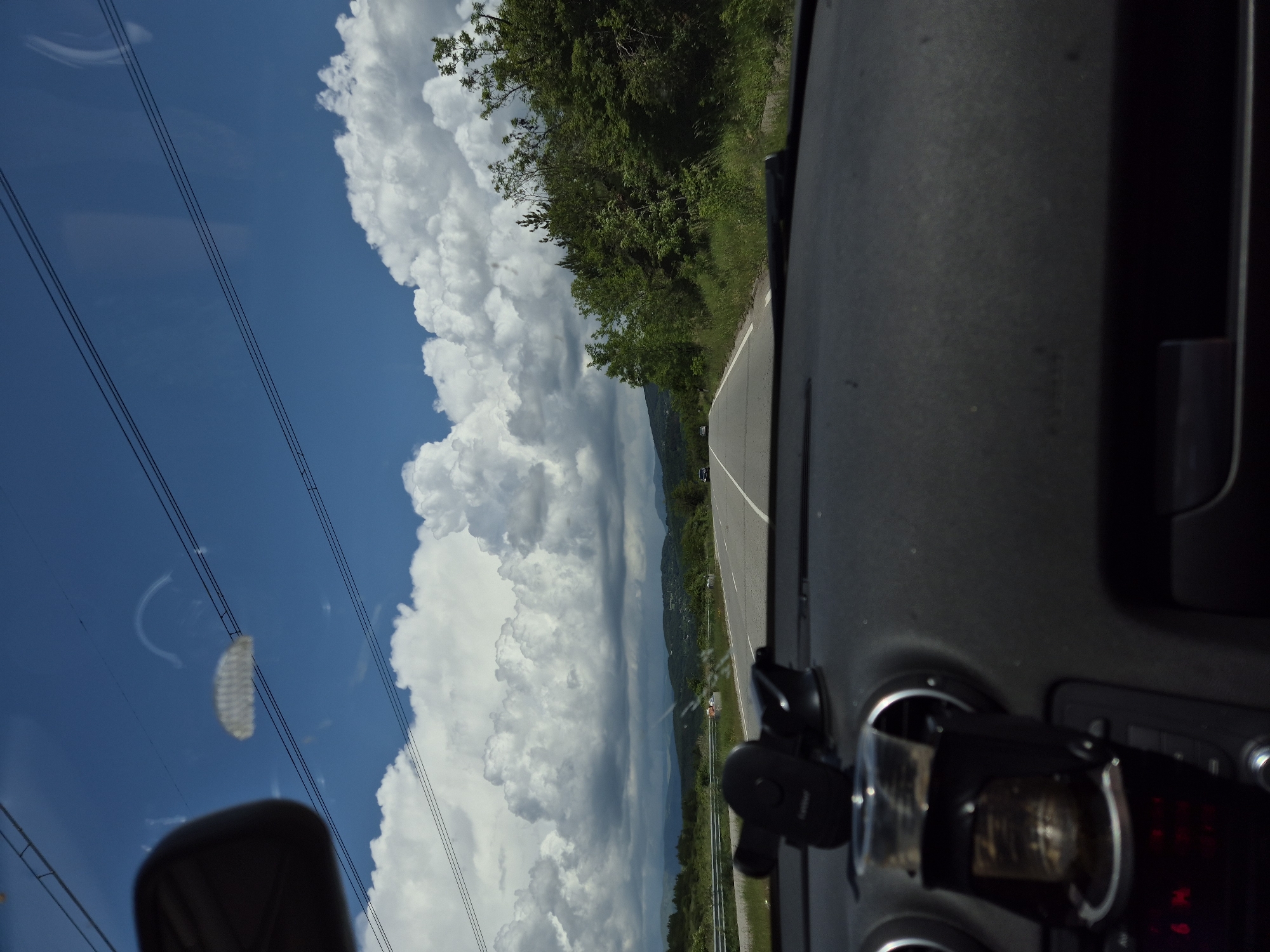 The image provides a daytime forward-facing view from inside a vehicle traveling in the area of Ponor, Bulgaria. The lower half of the frame displays the vehicle's dark gray dashboard. On the left side, a black car phone holder is mounted, and below it, a black cupholder, attached to an air vent, contains a translucent plastic cup with a small amount of residual liquid. A portion of the rearview mirror is visible in the upper left, reflecting interior elements of the vehicle. The windshield shows various reflections and smudges, including distinct horizontal power lines, a semicircular arc, and a grid-like pattern, superimposed against the sky.
The vehicle is on an asphalt road with a solid white line marking its right edge and a broken white line indicating lane separation. A silver metal guardrail runs parallel to the road on the left, beyond which is a vegetated slope. The immediate roadside is covered with green grass, trees, and shrubs.
In the mid-ground and background, the road extends into a rural and mountainous landscape. Distant green hills are densely covered with foliage, and other vehicles are faintly visible on the road ahead. The sky transitions from clear blue in the upper regions to being predominantly filled with large, white cumulus clouds, some of which appear darker and more diffused.