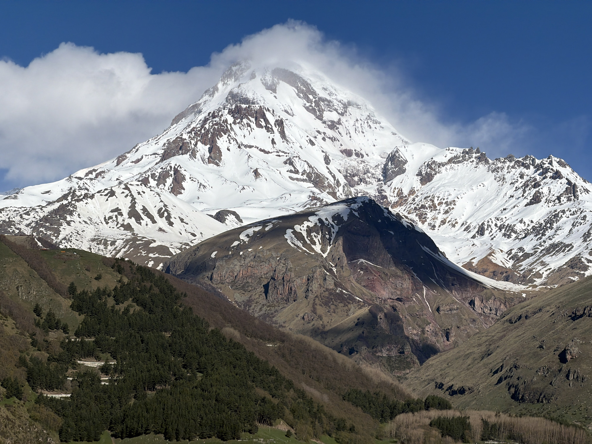 A daytime shot reveals Mount Kazbek, a stratovolcano, rising above the landscape near Stepantsminda, Georgia. The peak is snow-covered, with some areas of exposed rock. A cloud shrouds the very top of the mountain. Below the peak is a rocky mountain with exposed earth and some snow cover. Lower down, the landscape transitions to green hills with coniferous forests. At the very bottom of the frame, there are deciduous trees. A road with hairpin turns ascends one of the green hills. The sky is blue with scattered clouds.