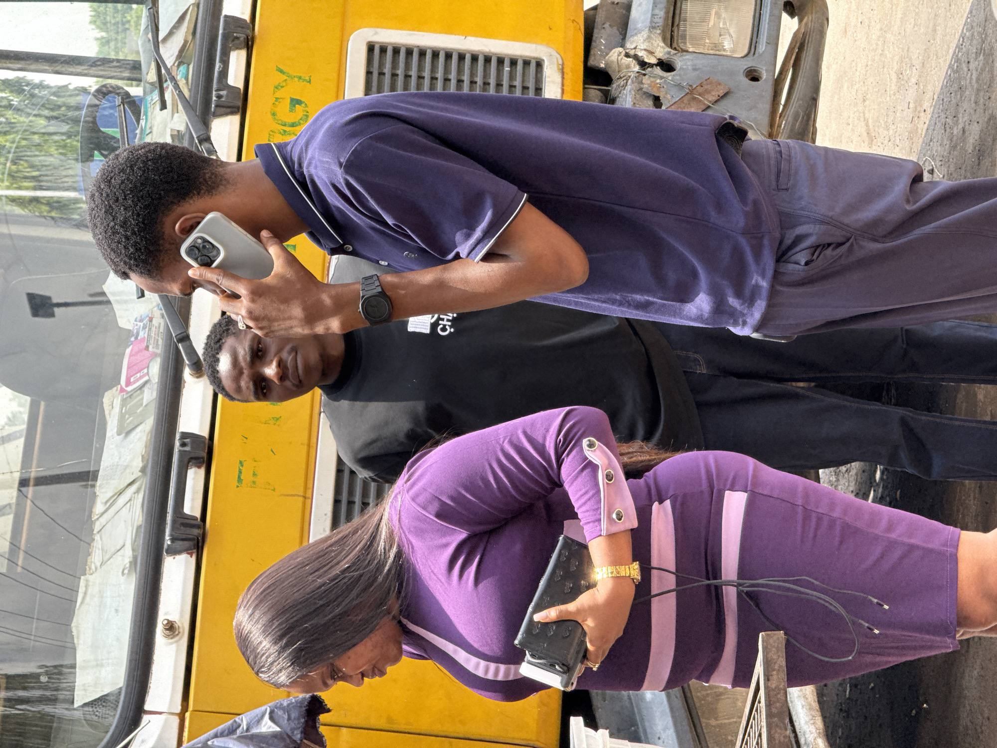 Three individuals stand alongside a yellow vehicle. A man in a navy blue polo and matching trousers is on the phone, while a man in a black t-shirt stands behind him. A woman in a purple dress holds a black wallet. The location is suggested to be Abuja, Nigeria.