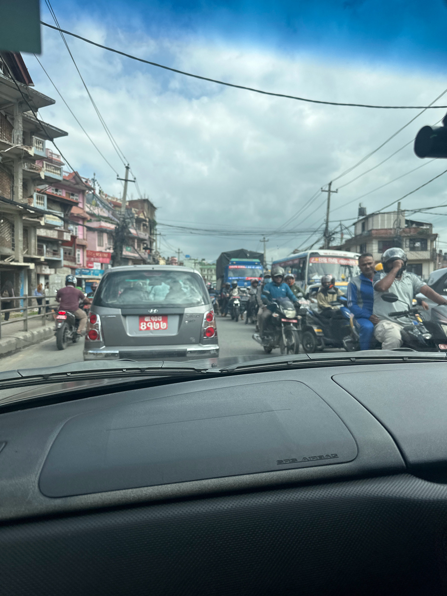 The image captures a busy street scene in Kathmandu, Nepal, with a view from inside a vehicle looking forward. The foreground is dominated by the interior of a car, including the dashboard with visible "SRS AIRBAG" text and a windshield wiper.
The street itself is filled with traffic, predominantly motorcycles and a few cars and buses. People on motorcycles are seen wearing helmets and casual clothing. A gray Hyundai sedan is prominent in the middle ground, with a red license plate. Buildings line both sides of the street, appearing to be multi-story structures with shops or residences. The sky above is partly cloudy. The overall impression is a typical, congested urban environment.