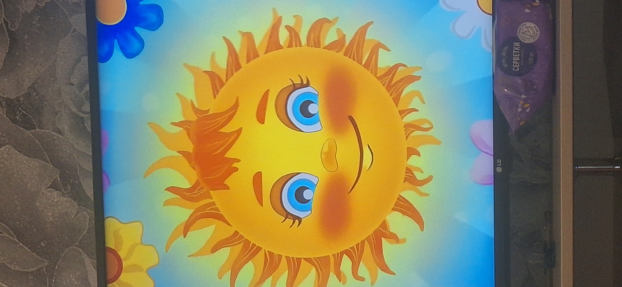 The image displays a cartoon sun with a smiling face and bright blue eyes, set against a blue sky with blurred floral elements. The sun has orange rays and rosy cheeks. Below the image on the television screen, a purple package of wet wipes with Russian text is visible. The television is placed on a wooden shelf. The background of the image shows a textured grey wall. There is no identifiable information in the image that links it to Bila Tserkva, Ukraine.