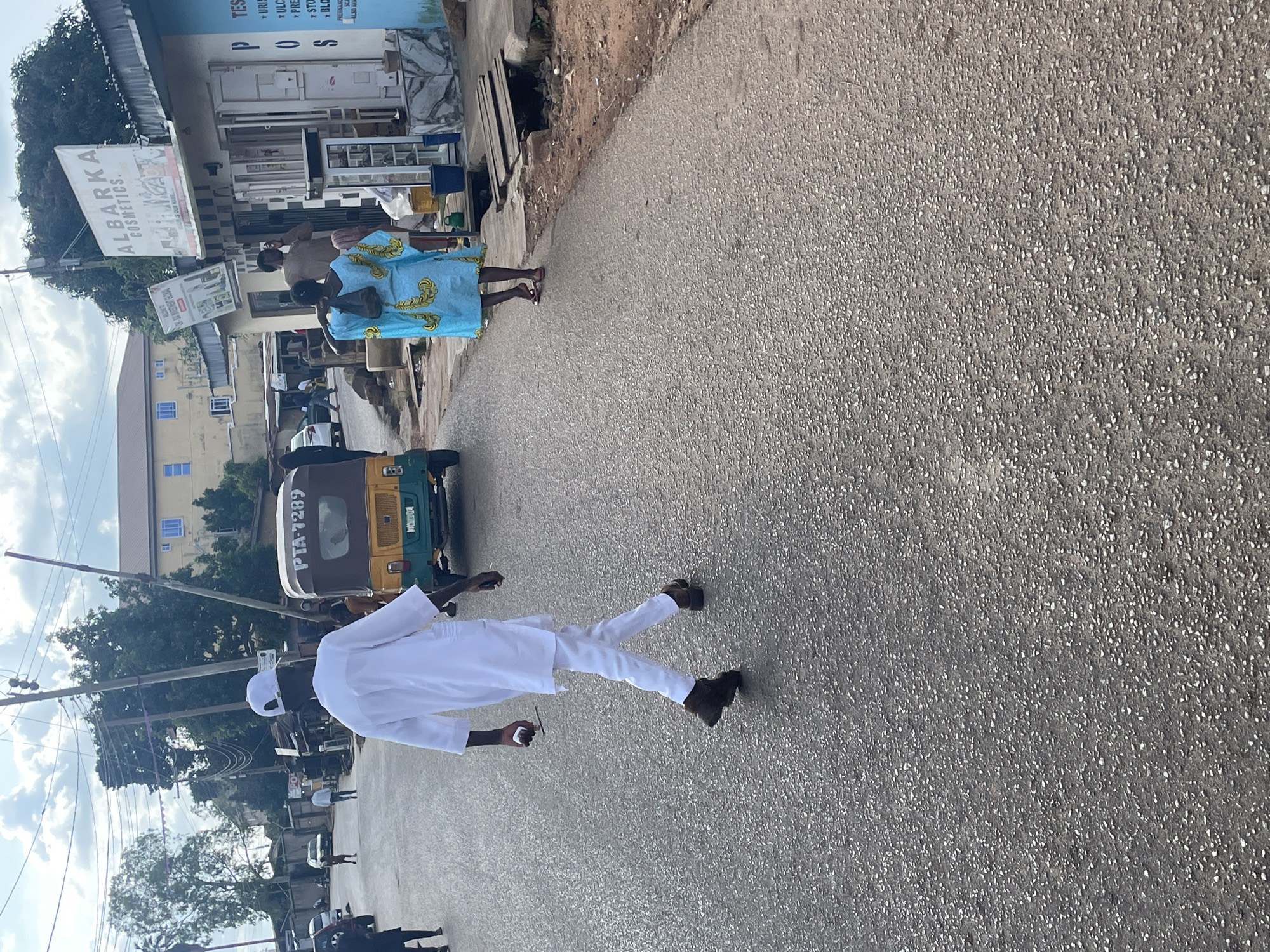 The image depicts a street scene in Jos, Nigeria, likely during the day under a cloudy sky. In the foreground, a man dressed in a white traditional outfit walks away from the camera, holding a white object in his right hand. To his left, a yellow and green auto-rickshaw, identified by the license plate "PTA 7289", is parked on the asphalt road. Further ahead, a woman in a vibrant blue and yellow patterned dress walks away from the camera, carrying a dark bag over her shoulder. Adjacent to the road, on the right side of the image, are several shops. One shop prominently displays a sign reading "ALBARKA COSMETICS". Another shop has signs with text such as "TEST", "PUR...", "ULC...", "PRE...", and "BLO...". The environment includes buildings, trees, utility poles with numerous wires, and other vehicles in the background, suggesting an urban or semi-urban setting. The road surface is asphalt with visible aggregate.