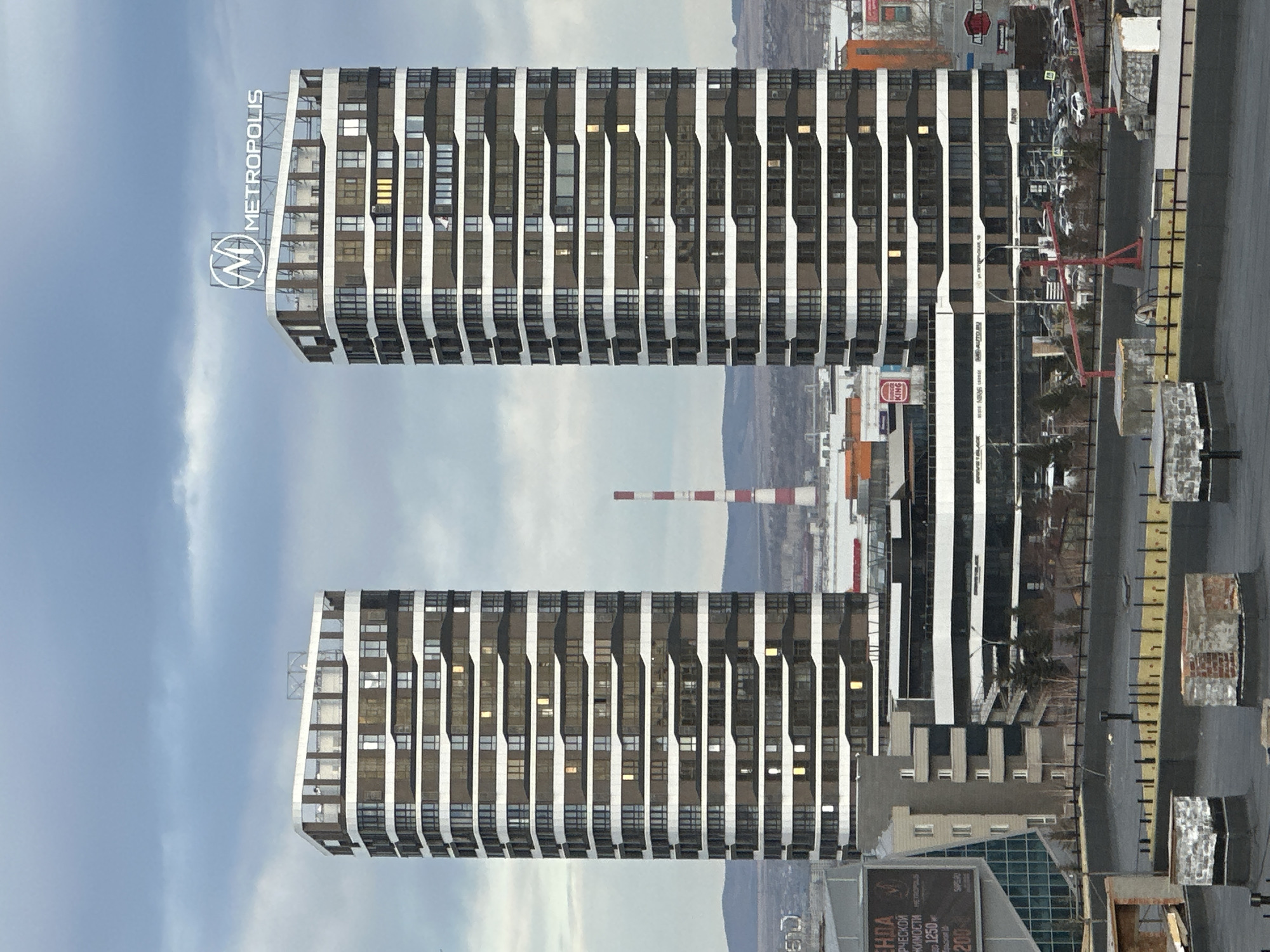 The image shows two high-rise buildings with a distinctive design of alternating dark and light vertical stripes, likely located in Krasnoyarsk, Russia, as "METROPOLIS" is visible on one of the buildings. The buildings have a modern architectural style. The surrounding environment includes other buildings, roads, and a distant mountainous landscape. A tall, red-and-white striped smokestack is visible in the background. In the foreground, there is a street with street infrastructure. The sky is partially cloudy. There's a construction site near the base of the tower.