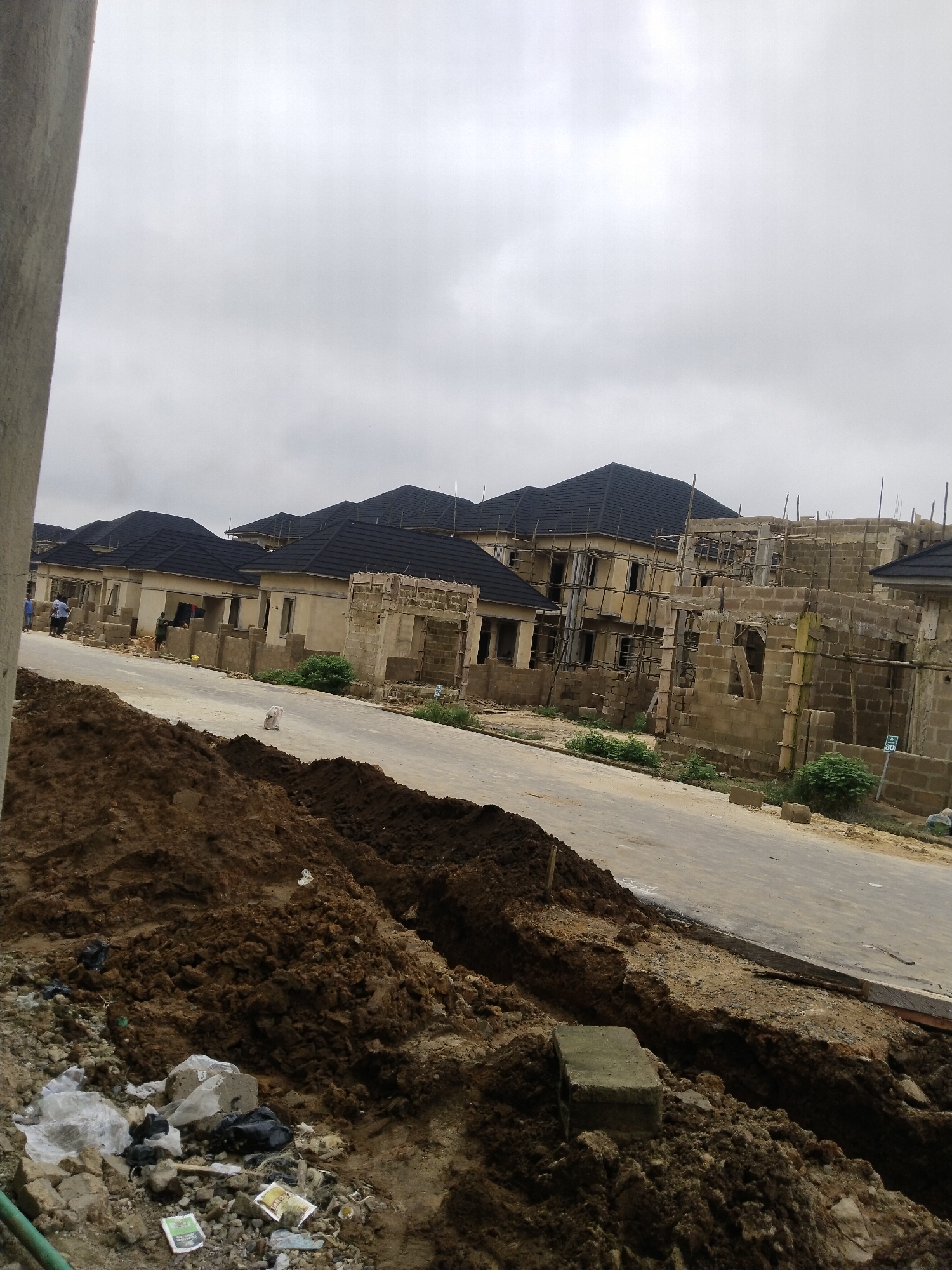 The image shows a street in a residential development under construction, with multiple houses in various stages of completion. The foreground features a pile of dark brown soil and debris, including plastic bags and scattered trash. A paved road runs through the scene, with a trench dug alongside it. In the midground, several houses with dark tiled roofs are visible. Some of these houses appear to be finished with exterior walls and windows, while others are still in the early stages of construction, with exposed brickwork and scaffolding. A few individuals can be seen walking on the road in the distance. The sky is overcast and gray. The overall impression is one of ongoing development in a suburban or semi-rural setting.