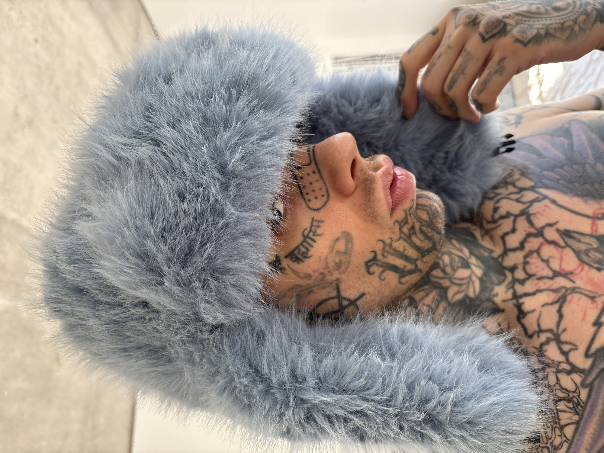 A pale-skinned person is lying down, their head adorned with a fluffy, light blue hat. The person is heavily tattooed on their face, neck, chest, and hands. The tattoos appear to be a mix of script, geometric designs, and floral motifs. The individual is lying on a light-colored surface, possibly a bed or a sofa. The location is unknown, and there is no indication of it being in Buenos Aires, Argentina.