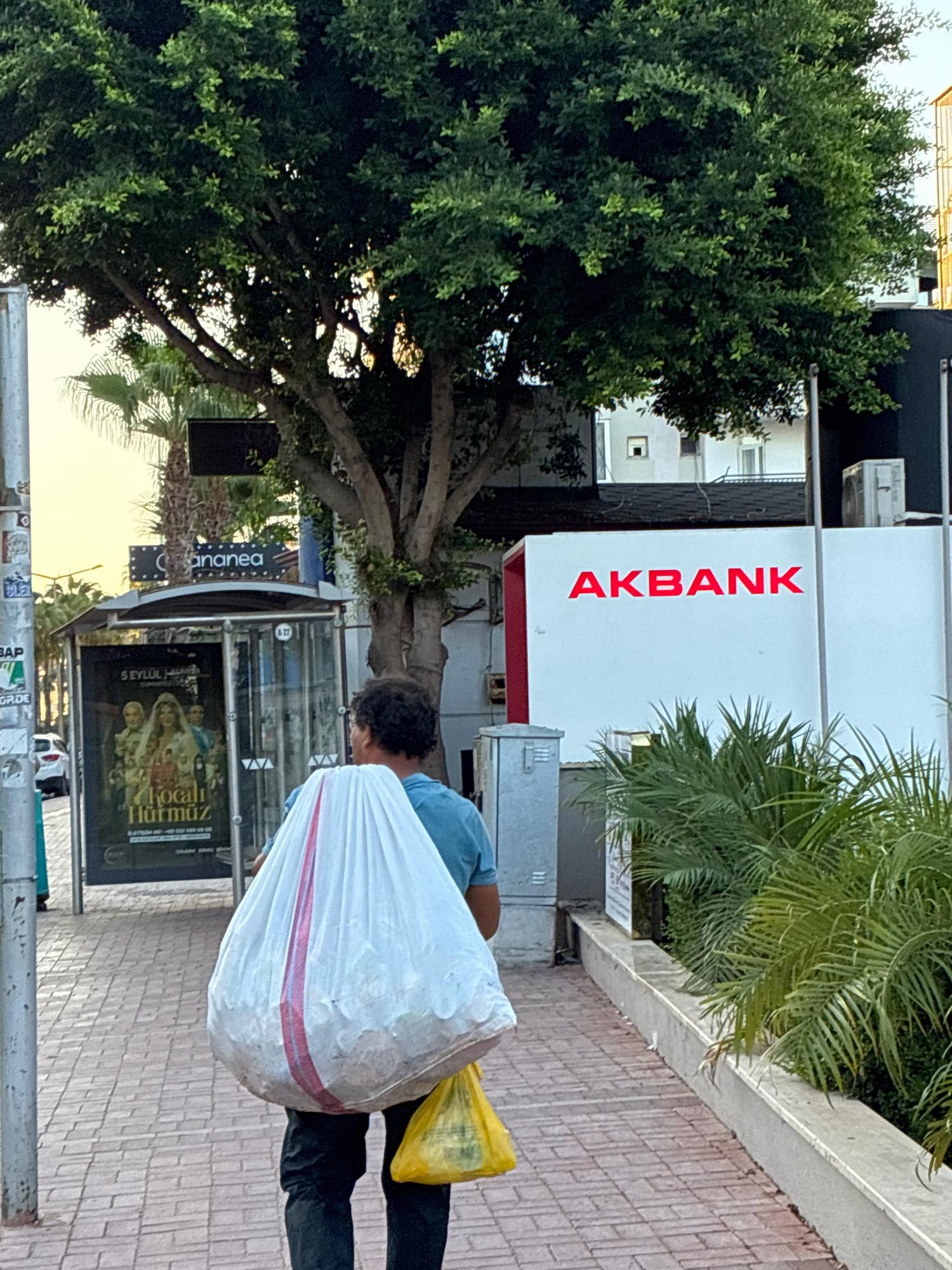 A man walks along a tiled sidewalk in Alanya, Turkey, carrying a large white plastic bag filled with what appears to be trash or recyclables. He also has a smaller yellow plastic bag in his right hand. He is wearing a blue t-shirt and dark pants. In the background, a building with the red "AKBANK" logo is visible, along with a bus stop shelter displaying a poster. Palm trees and other foliage are present, suggesting a warm climate. The sky is bright, indicating daytime. The scene captures a moment of daily life in the city.