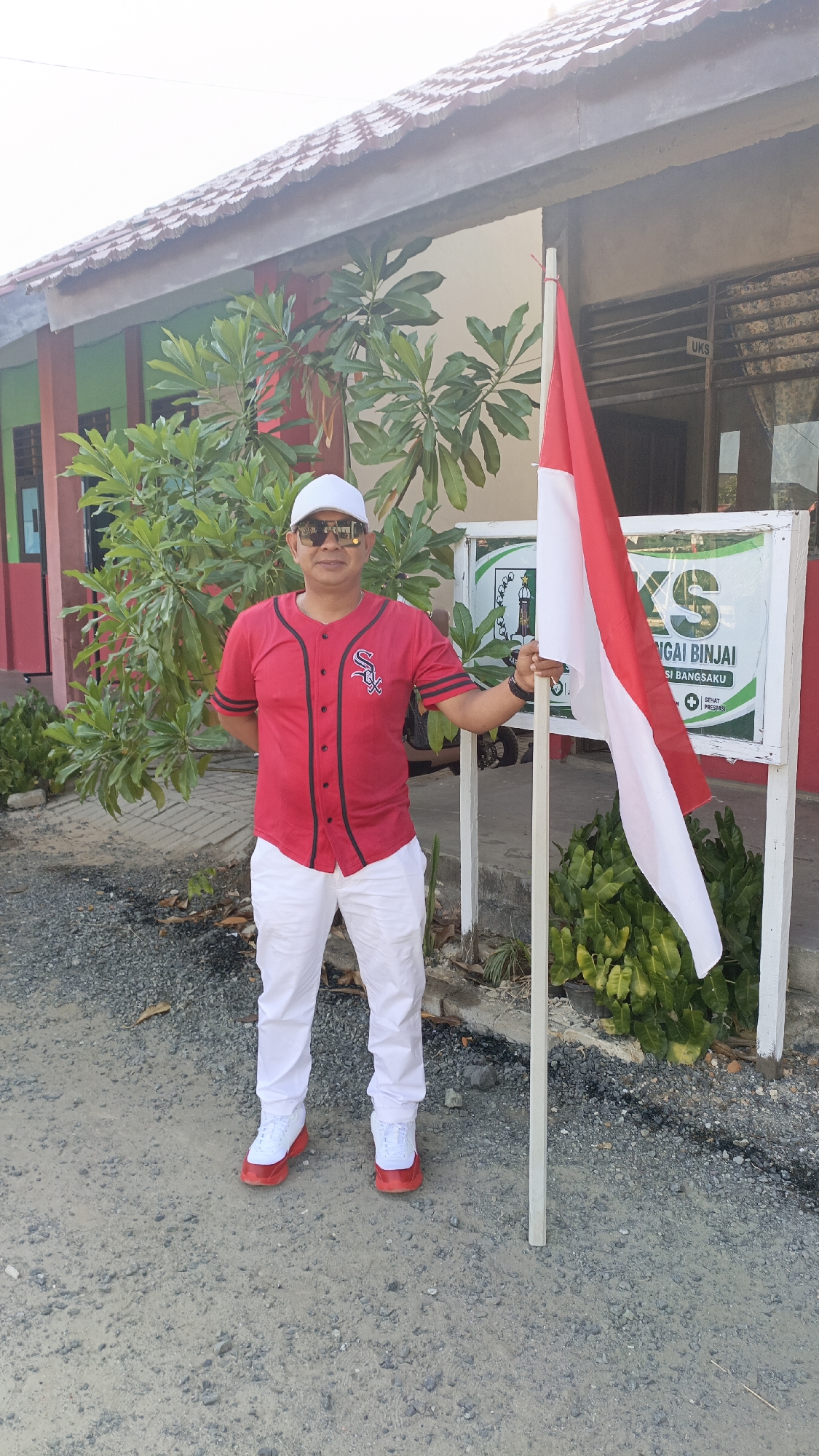 In Saring Sungai Binjai, Indonesia, a man stands outdoors holding a white flag pole with the Indonesian flag attached. He is wearing a white baseball cap, sunglasses, a red baseball jersey with black trim and a white logo on the left chest, white pants, and white sneakers with red accents. He is positioned in front of a building with a tiled roof and a green and red facade. A tree with large green leaves is to his left, and a white sign with green lettering is behind him and to his right. The ground is covered with gravel. The man is facing the camera and appears to be posing.