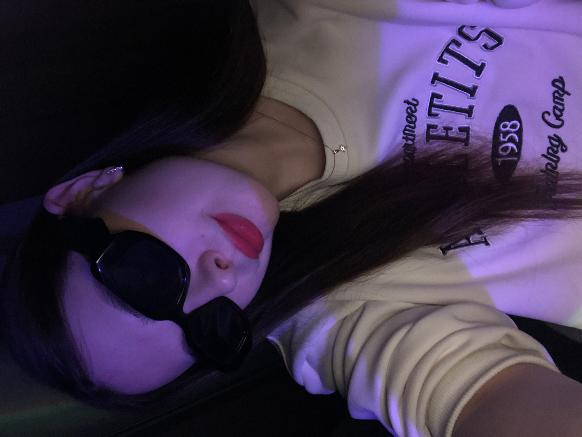 A woman with dark hair wearing dark sunglasses and red lipstick is lying down. She is wearing a light-colored sweatshirt with the text "ELITE 1958 Training Camp". She also has a small gold necklace and earrings. The lighting is purplish.
There is no indication that this image was taken in Noviy, Russia.