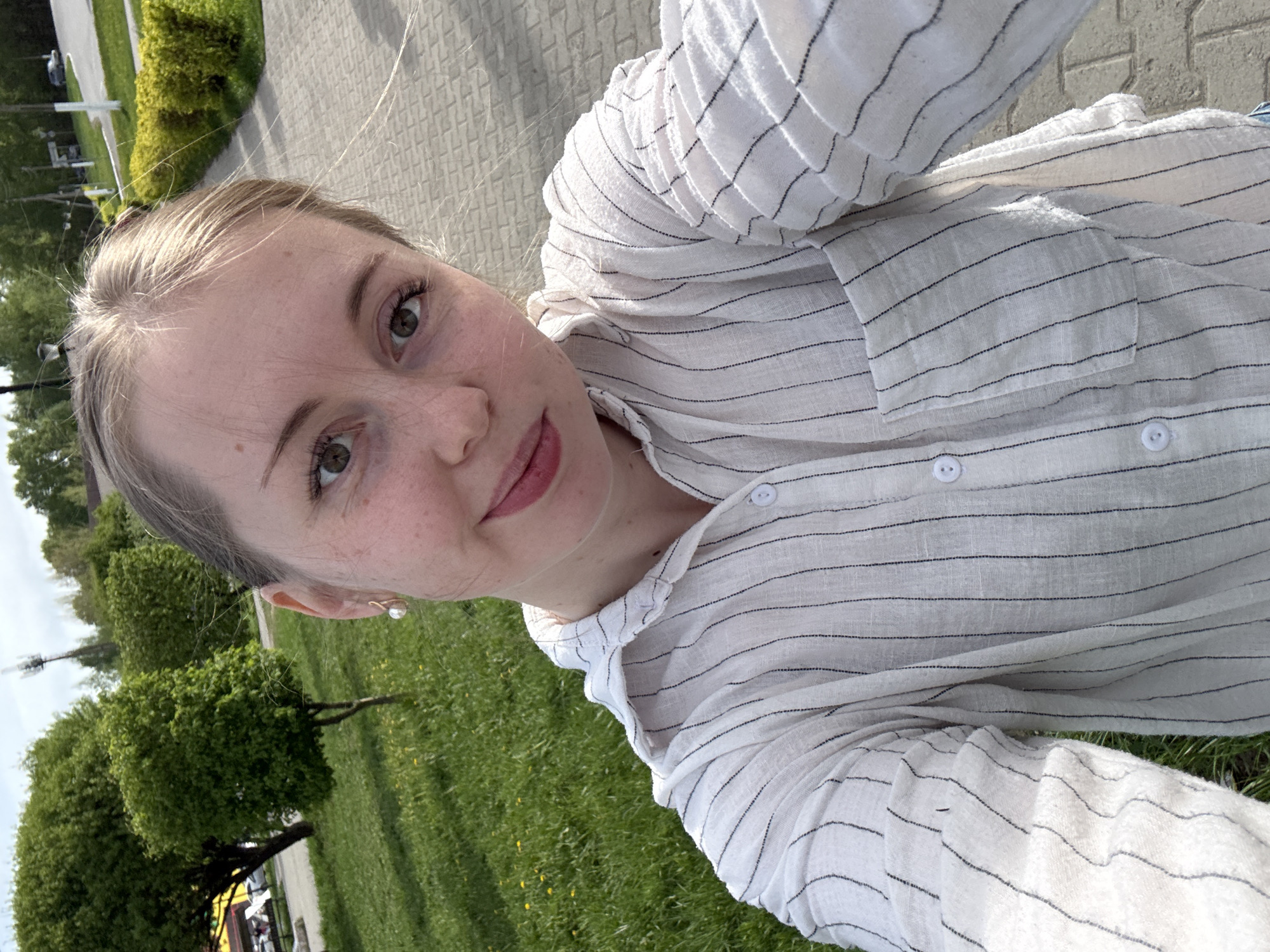 A selfie of a fair-skinned young woman with light brown hair and green eyes, wearing a white and black striped button-down shirt. Her head is tilted, and she is looking directly at the camera with a slight smile. The background consists of a city park, featuring green grass, trees, and a brick sidewalk or paved area. In the distance, there are streetlights and what appears to be a parking area with vehicles. The location is Unknown, Unknown.