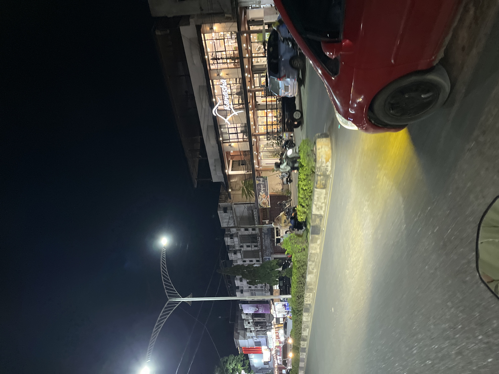 The image captures a nighttime street scene in Langsa, Indonesia, featuring a prominent, well-lit modern building with the name "Ranasha" displayed in illuminated script. The building appears to be a restaurant or commercial establishment with large glass windows revealing a warmly lit interior. Several vehicles, including cars and motorcycles, are present on the road and parked along the street. A red car is in the foreground, partially obscuring the view of the street. The street is illuminated by streetlights and the lights from the businesses. The environment suggests an urban setting with sidewalks, landscaping, and other buildings lining the street. The sky is dark, indicating it is night.