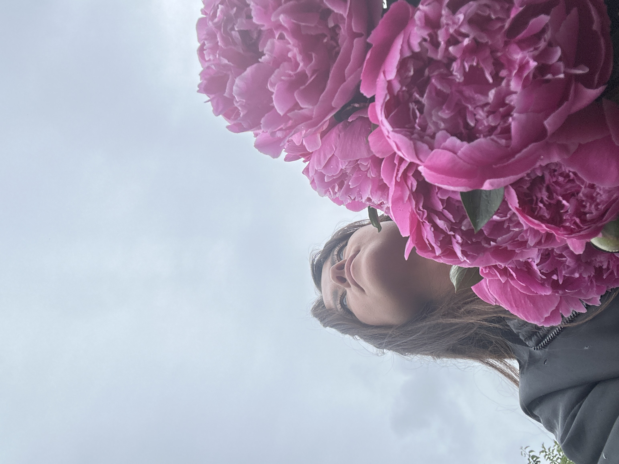 A low-angle, outdoor shot captures a woman looking up at a bouquet of bright pink peonies against a gray, overcast sky. The woman, positioned on the left side of the frame, has dark hair and is wearing a dark jacket. The peonies dominate the foreground and right side of the image, their petals detailed and layered. The background consists of a uniform, cloudy sky, with no discernible cityscape or specific location in Chaikovsky, Russia.