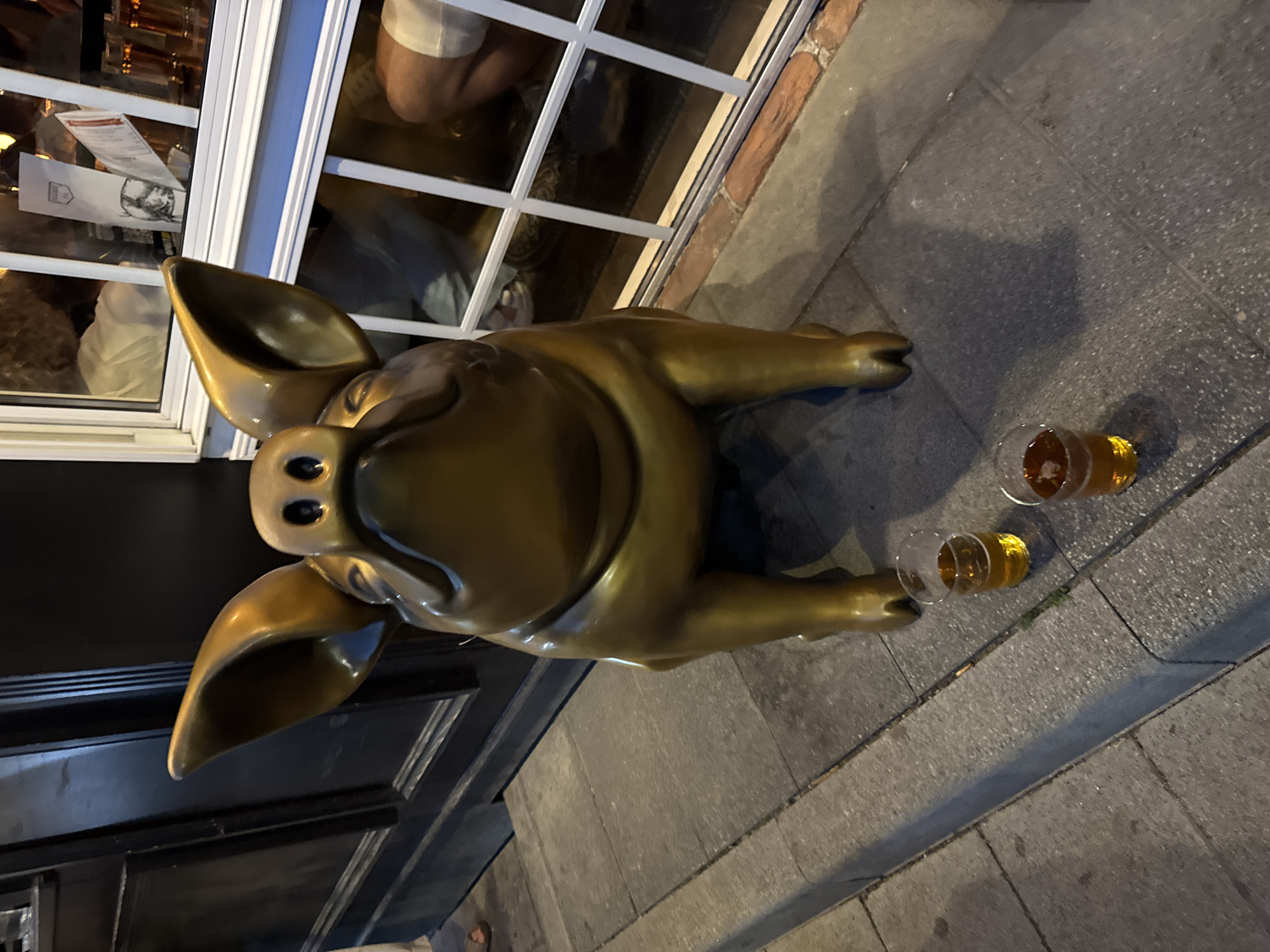 The image depicts a large, golden bronze statue of a pig, positioned outdoors on a sidewalk. The pig statue is centrally located and appears to be the primary subject. In front of the statue, on the pavement, are two clear plastic cups containing amber liquid, presumably beer.
Behind the statue is a building facade with dark wooden paneling on the left and a large window with white cross-panes on the right. Through the window, the interior of what appears to be a bar or restaurant is visible. People can be seen inside, and there are some papers and menus displayed. The lighting suggests it is either dusk or nighttime, with some artificial light sources contributing to the scene. The ground is paved with stone tiles, and a concrete curb separates the sidewalk from the street. The overall scene is a street-level view of the pig statue and its surroundings.
This specific location is not in Moscow, Russia, but rather appears to be a public space, possibly a street or plaza outside a commercial establishment.