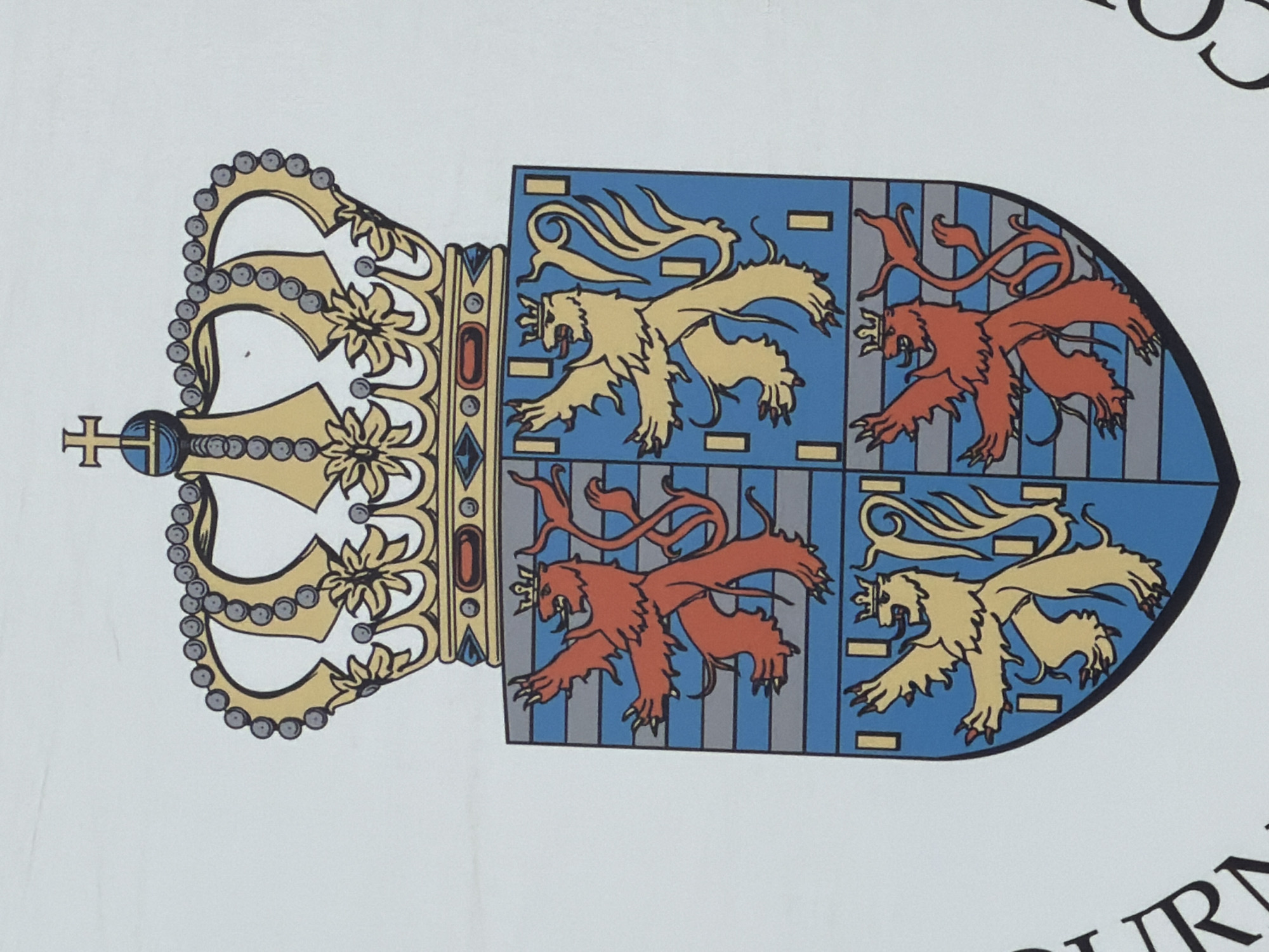 The image displays a close-up view of a coat of arms, likely a national or regional emblem. The coat of arms features a large, ornate golden crown atop a shield. The shield is divided into four quarters, each containing a rampant lion. The lions alternate in color between red and yellow, and the background of each quarter is a pattern of blue and grey horizontal stripes with thin vertical grey lines. Some of the lions are depicted wearing smaller crowns.
To the left of the coat of arms, partially visible, is text that appears to be "URAN". To the right, also partially visible, are decorative scroll-like elements, possibly part of a larger design.
The background of the image is a plain white surface.
While the image itself does not provide specific location details, the mention of Leudelange, Luxembourg, suggests this coat of arms is associated with that municipality. If this is indeed the coat of arms of Leudelange, it would likely be found on public buildings, official signage, or historical markers within the commune of Leudelange.