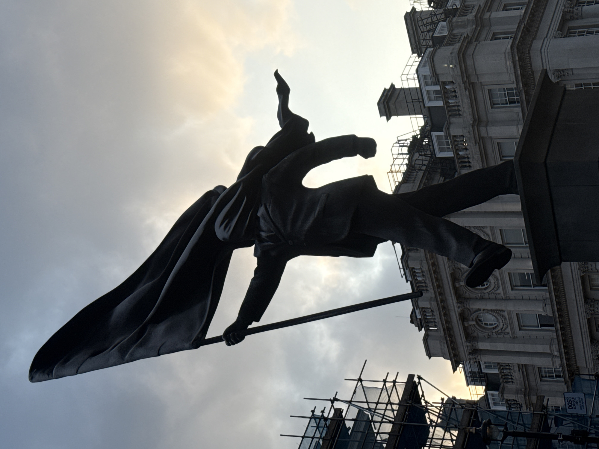 The image is a low-angle, upward-tilted shot of a dark statue against a cloudy sky in an urban setting. The statue depicts a figure in what appears to be a cloak or cape, with one arm raised and holding a pole or staff. The background includes a building with architectural details such as windows, ledges, and decorative elements. Scaffolding is visible near the bottom of the frame. The statue is likely a monument or public artwork situated in London, United Kingdom.