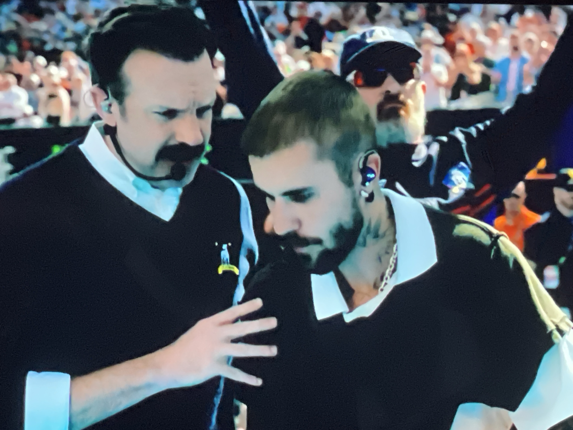 Two men are in the foreground of a blurred crowd. The man on the left, facing right, wears a black sweater vest over a white collared shirt and has a mustache and earpiece. The man on the right, facing left, wears a black shirt with a white collar and has an earpiece in his right ear and tattoos on his neck. His left hand is on the chest of the man to his left. In the background, a man with a beard, sunglasses, and a blue baseball cap is visible.