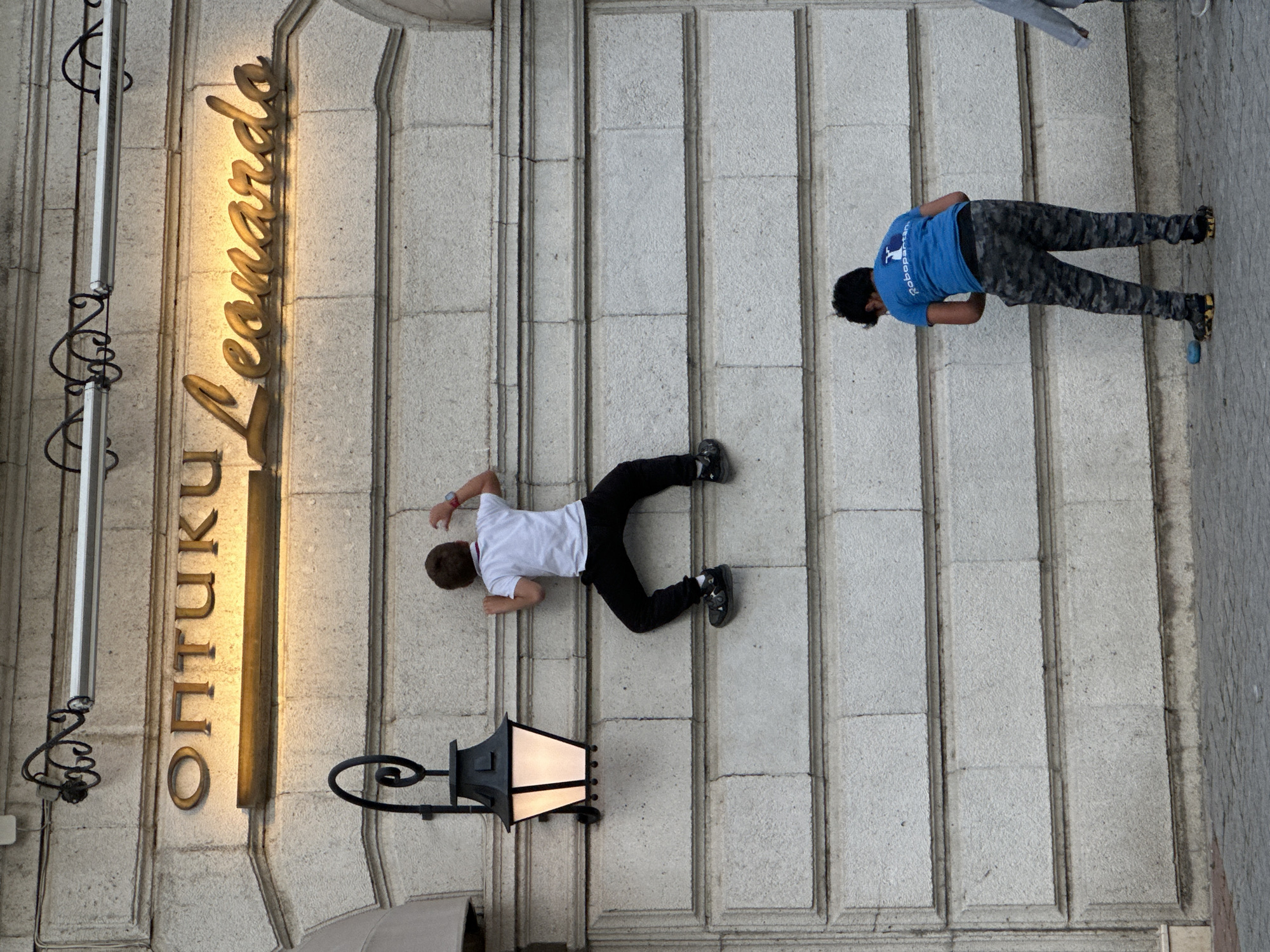 The image shows two people lying on a stone staircase outside a building. A white sign with the name “Оптики Leonardo” in gold Cyrillic letters is on the building’s facade. A black ornate lamppost is also visible near one of the people. The person closer to the sign is wearing a white shirt and black pants and shoes. The other person, located further up the staircase, is wearing a blue shirt and grey patterned pants. The location is in Varna, Bulgaria, as implied by the sign language (Cyrillic).