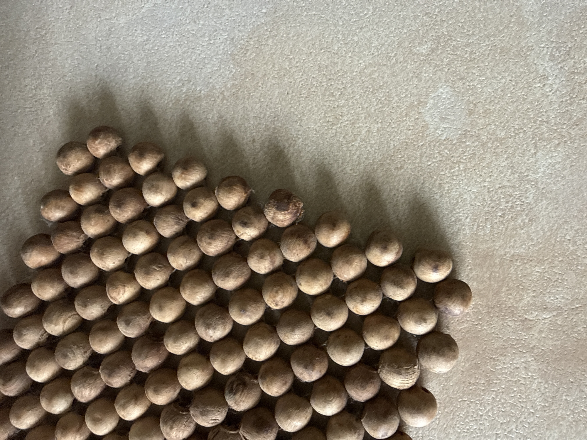 The image displays a close-up, high-angle view of numerous small, spherical objects, likely seeds, nuts, or wooden beads, arranged in a dense, roughly triangular formation. These objects, predominantly in shades of light brown and beige, possess a slightly textured, dull surface and are uniform in size. They are tightly packed in rows, creating a pattern that fills the lower-left portion of the frame and tapers towards the upper-right.
The objects rest upon a light-colored, granular surface, possibly concrete, stucco, or a coarse fabric, which is a consistent beige or light tan. Shadows are cast by each object onto this background, indicating a light source from above and slightly to the left, adding depth and highlighting their three-dimensional nature.
No specific activity or event is taking place; the objects are static, arranged in what appears to be an intentional display or pattern. There are no people, animals, or visible text. The lighting suggests daytime. This scene is situated in Vinnytsia, Ukraine.