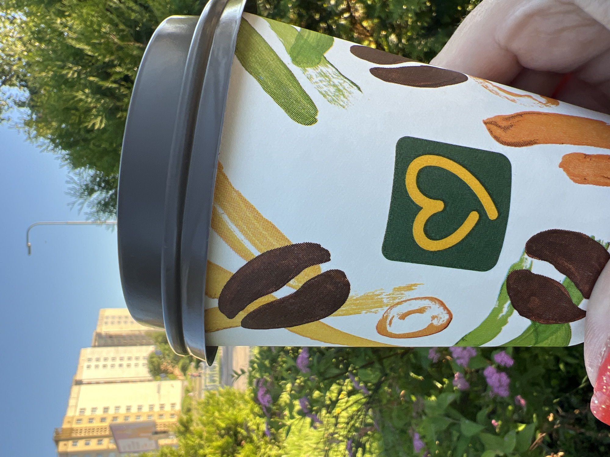 In Minsk, Belarus, a close-up shot focuses on a disposable coffee cup with a dark lid, held in a hand with red-painted fingernails. The cup features a playful design of brown coffee beans, green brush strokes, and orange accents. Prominently displayed on the cup is a yellow heart symbol within a green square. The background reveals a blurred urban landscape with tall, light-colored apartment buildings under a clear blue sky. Lush green trees and bushes, some with small purple flowers, frame the cup, suggesting an outdoor setting with natural elements juxtaposed against the city infrastructure. The sunlight casts a warm glow on the scene.
