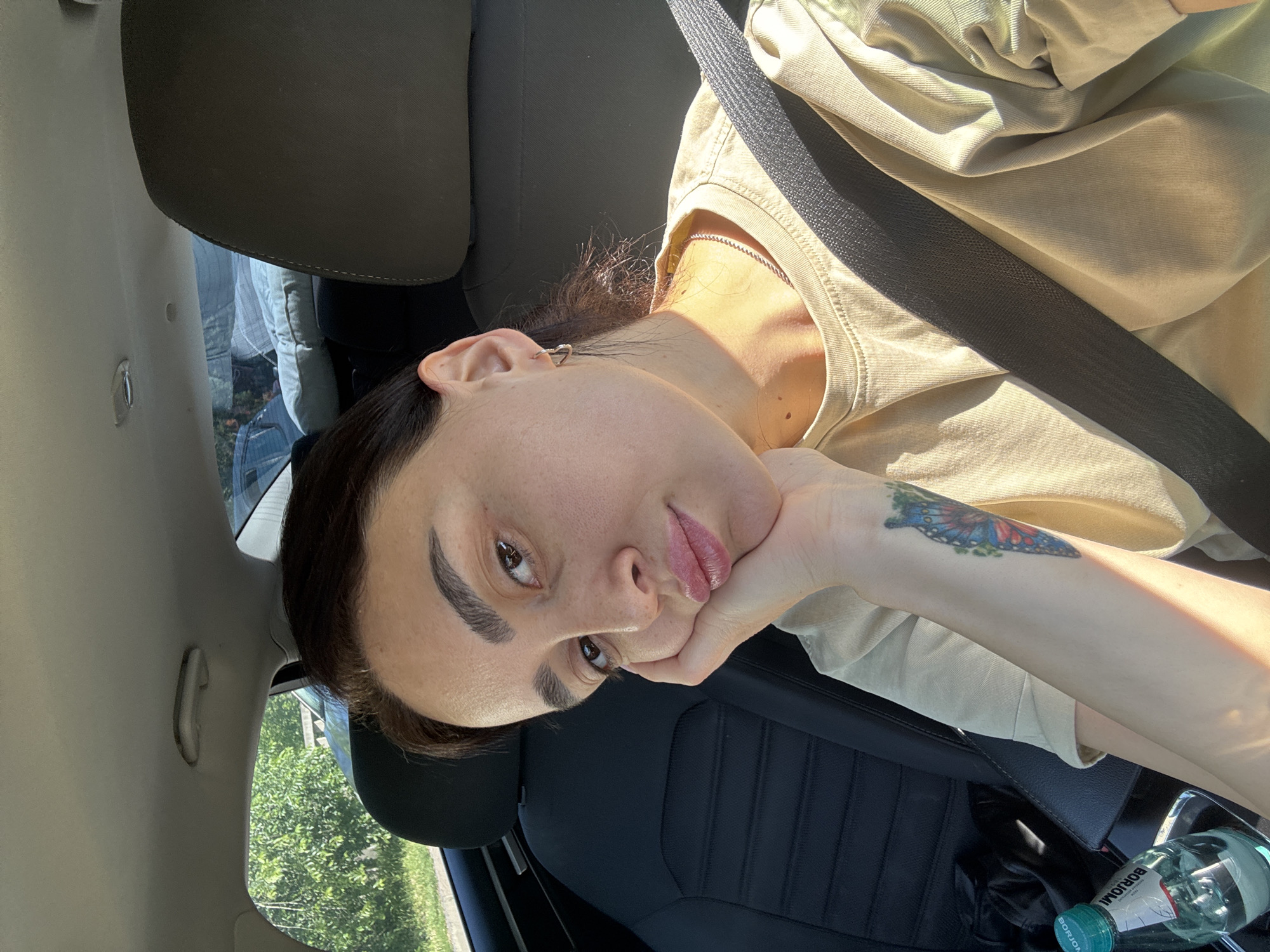 A woman is seated in the driver's seat of a car, wearing a beige t-shirt and a seatbelt. She rests her head on her hand, which has a butterfly tattoo. A bottle of Borjomi mineral water is visible in the car. The environment visible through the window suggests the presence of greenery, possibly urban trees. Given the brand of water, the location is likely Tbilisi, Georgia.