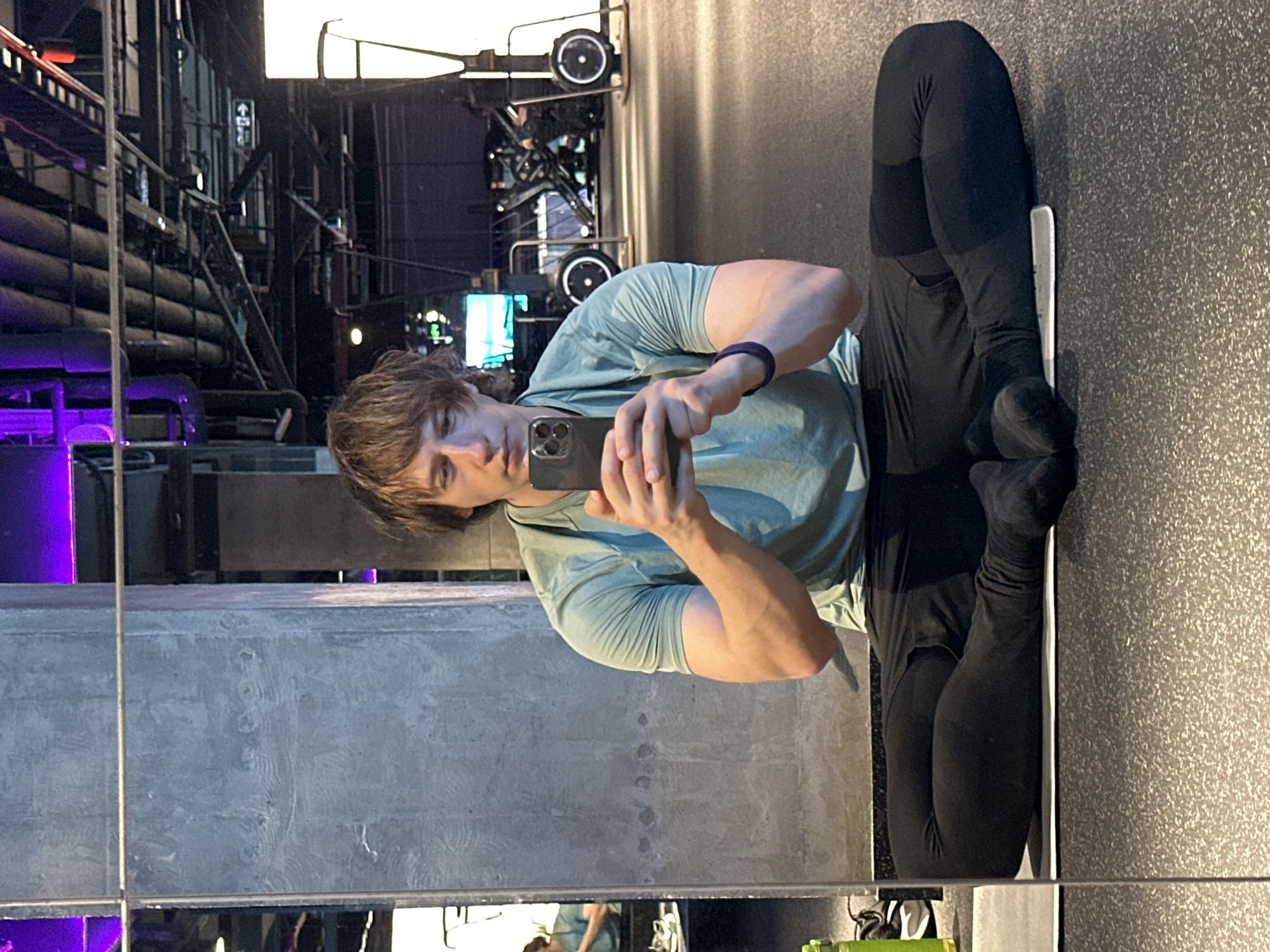 The media file is a vertical, full-body mirror selfie taken indoors. The main subject is a young person with light brown hair, wearing a light green t-shirt and black leggings with black socks. They are holding a smartphone in their right hand, angled to capture the reflection. The person's left arm is bent, resting on their leg, and a dark purple fitness tracker is visible on their wrist. They appear to be in a gym or fitness studio, as indicated by the textured floor and the blurry background which suggests gym equipment or industrial elements. The lighting is dim, with some purple and white lights visible in the background. The reflection also shows a sliver of another person in the lower part of the frame. The overall mood appears casual and focused on the individual. There is no visible text in the image.