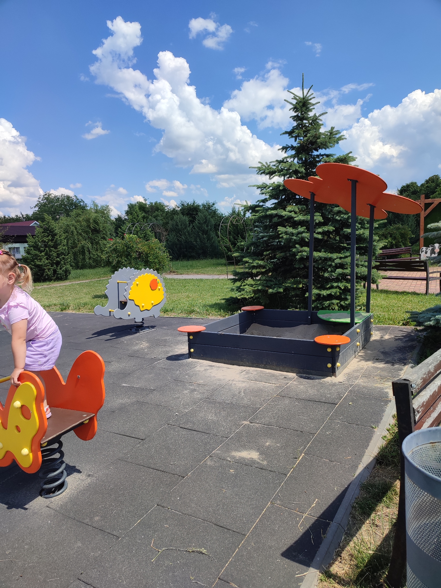 This is a full, eye-level, outdoor shot of a children's playground on a sunny day. The playground has rubberized flooring and is equipped with a sandbox, a seesaw, and a spring rider. In the foreground, a young girl with blonde pigtails, wearing a purple t-shirt and shorts, is holding onto the back of an orange and yellow butterfly-shaped spring rider. Behind her, a gray and yellow hedgehog-shaped seesaw is visible. The sandbox, a dark blue rectangular structure, is in the midground to the right, with an orange canopy overhead. A large fir tree stands behind the sandbox. In the background, there are green trees, a building with a red roof, a wooden bench, and a trash can. The sky is blue with fluffy white clouds. The location is identified as a playground in the city of Voskobiinyky, Ukraine.