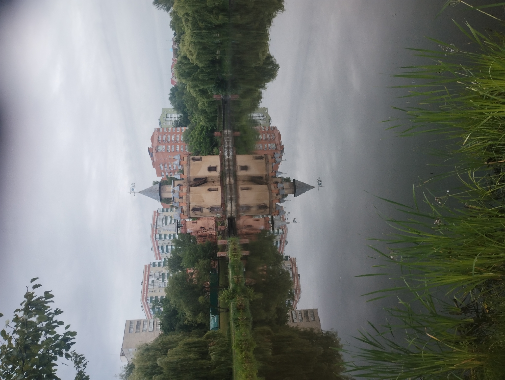The image shows a picturesque scene in the city of Sumy, Ukraine, featuring a small, fairytale-like castle situated on the bank of a tranquil lake. The castle, with its turreted roof and sandy-colored walls, is reflected in the still water, creating a symmetrical and captivating view. In the background, modern apartment buildings rise above lush green trees, contrasting with the historic charm of the castle. The sky is overcast, casting a soft, diffused light over the landscape. The foreground is occupied by tall reeds growing along the edge of the lake, adding a natural, wild element to the composition. The scene is devoid of any people or discernible activity, presenting a serene and peaceful ambiance.