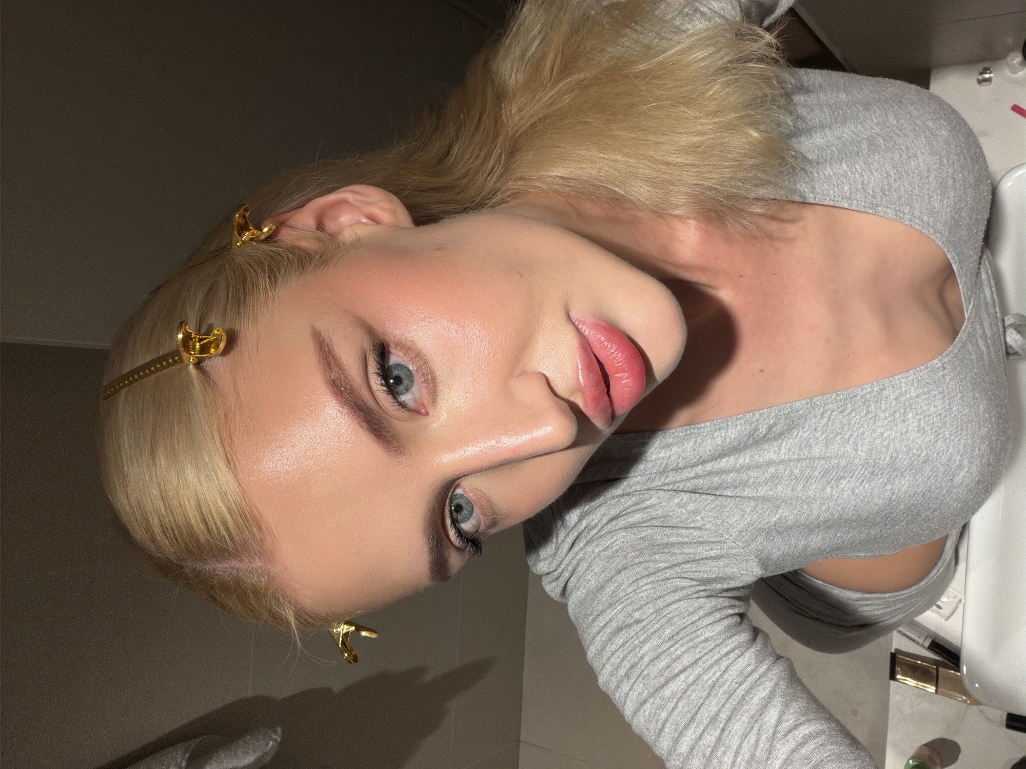A fair-skinned blonde woman with light blue eyes is pictured indoors, likely in a bathroom. She is wearing a light gray, long-sleeved, ribbed top. Her blonde hair is held back with gold-colored hair clips. She has makeup on, including blush and lip gloss. The background is dark, but a sink and some makeup products are visible. The location in the city of Belgrade, Serbia, cannot be determined from the visual information provided.
