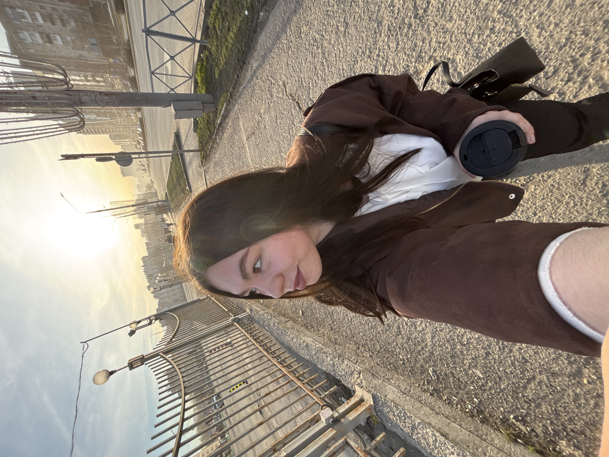 A selfie taken on a city street in Perm, Russia, at sunset. A woman is the primary subject, visible from the right side of her face and shoulders up. She has long, dark hair and is wearing a brown jacket over a white top. Her left hand holds a black travel mug. A black leather bag is partially visible behind her. The scene is a city street, with a metal guardrail running along the bottom left side. In the background, there's a road, buildings, street lamps, and a glimpse of the city skyline bathed in the golden light of the setting sun. The setting appears to be a sidewalk adjacent to a street with sparse vehicular traffic. The pavement is light gray and shows signs of wear.