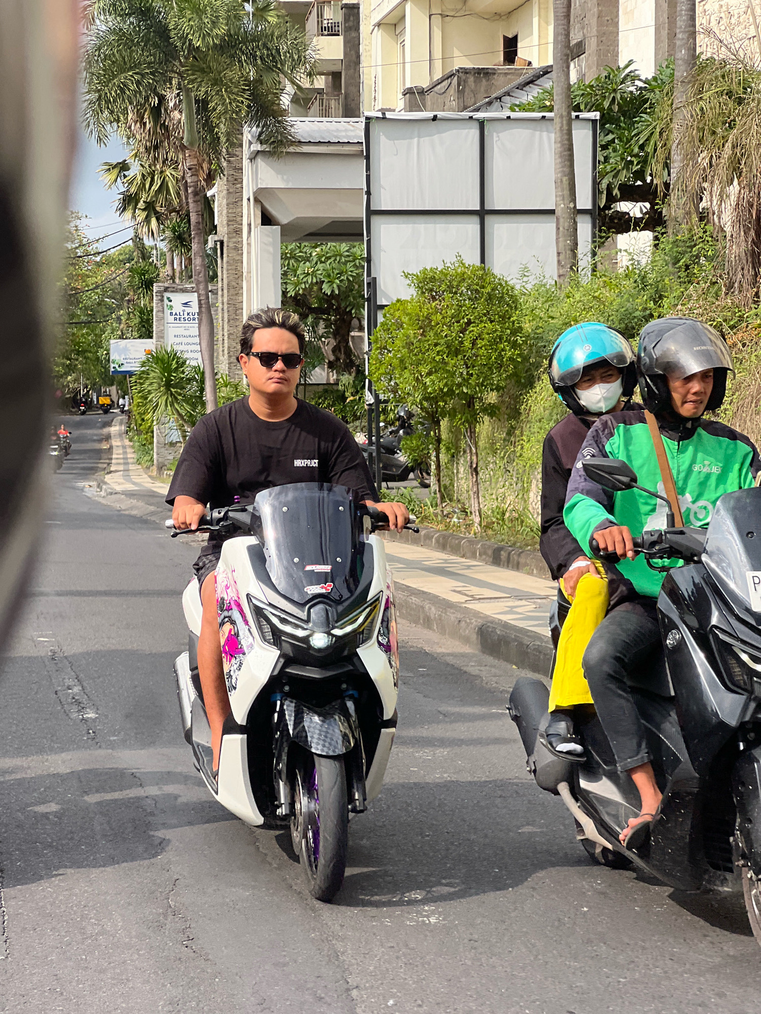 In Kuta, Indonesia, a man with styled blonde-tipped hair and sunglasses rides a white and pink scooter, wearing a black t-shirt with "HRXPRCT" printed on it and patterned shorts. He is on a paved road with a sidewalk to his left, lined with tropical trees and buildings. Behind him, another scooter carries two passengers. The person in front wears a turquoise helmet and a white face mask. The person behind them wears a dark helmet and a green jacket with "GOJEK" branding, black pants, and yellow leg coverings. The Gojek rider is looking to the right. The background shows a building with multiple balconies and lush greenery. Sunlight casts shadows on the road, indicating a bright day.