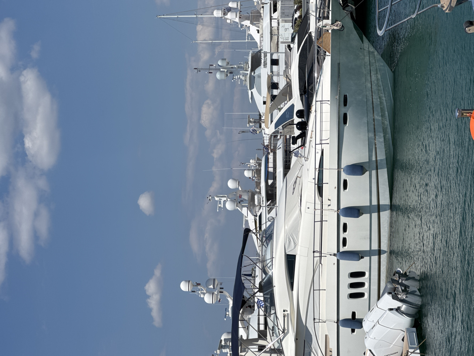 In the city of Moschato, Greece, a fleet of white yachts is docked in a harbor under a clear blue sky with scattered white clouds. The foreground features the stern of a large white yacht with outboard motors. Behind it, a larger white yacht is prominently positioned, its hull partially covered by a white tarp. This yacht has dark blue fenders hanging along its side, and several rectangular windows are visible. Further back, other white yachts of varying sizes are docked, some with masts and radar equipment. The water in the harbor is a deep teal color, with ripples reflecting the bright sunlight. The scene is peaceful, with no people clearly visible or actively engaged in any action, suggesting a quiet day at the marina.