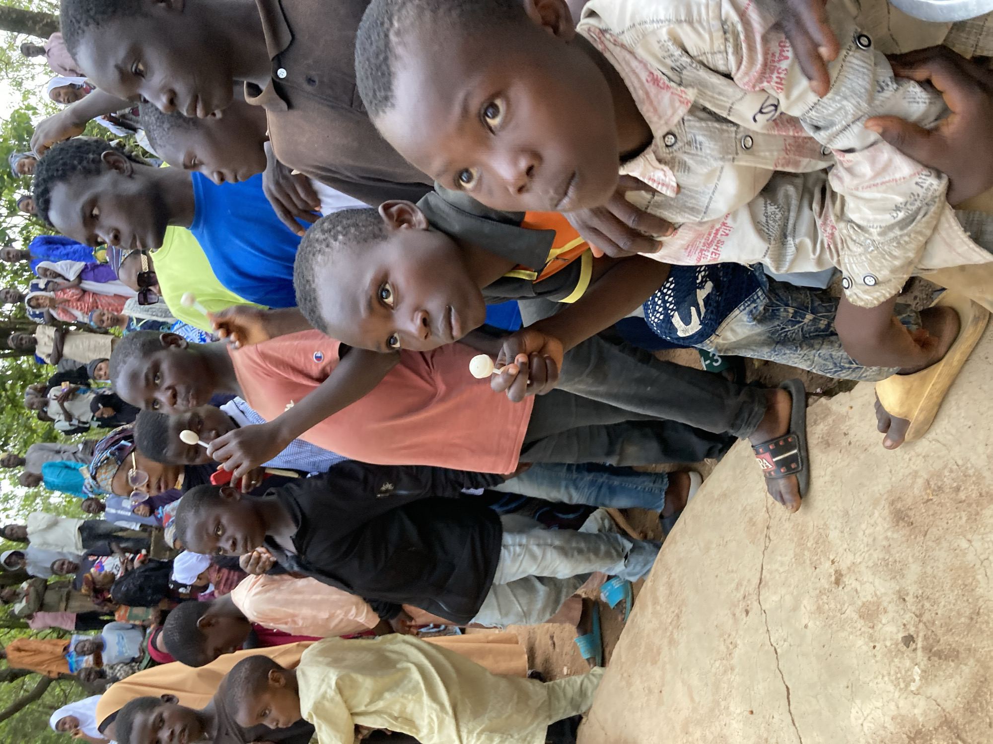 The image depicts a group of people, primarily children, gathered outdoors in what appears to be a semi-urban or rural setting. The ground is covered in dirt and rough concrete, with some vegetation visible in the background. The people are dressed in a variety of casual and traditional clothing, including robes, shirts, and head coverings. Many of the children are looking in the same direction, with some holding lollipops. The overall atmosphere suggests a community gathering or event. The provided location is the city of Jos, Nigeria.