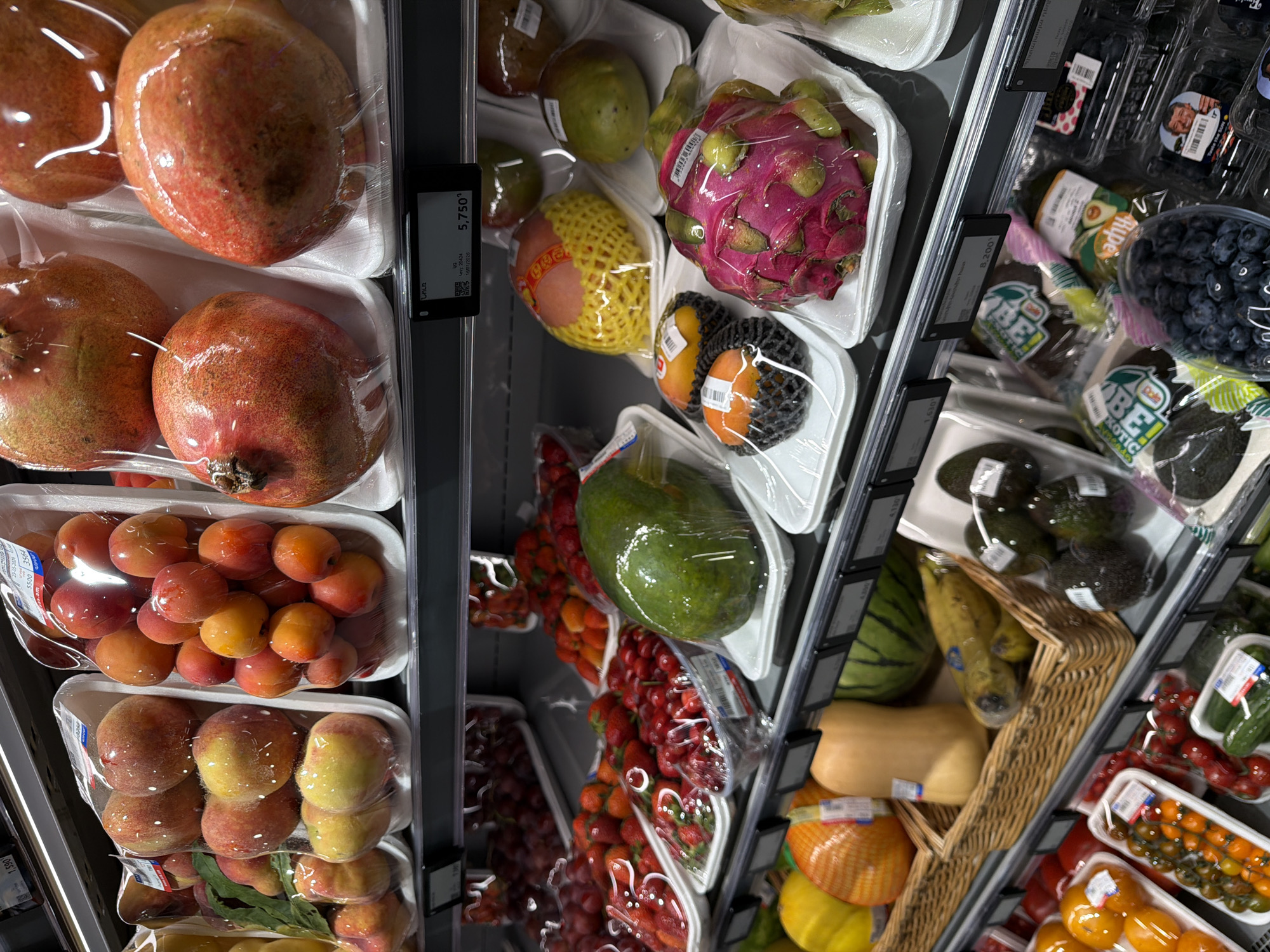 This image shows a shelf in a store, probably in Yerevan, Armenia, due to the presence of Armenian script on a price tag (Նուռ). The shelf is stocked with a variety of fruits and vegetables, many of which are packaged in plastic wrap on white styrofoam trays.
Fruits visible include pomegranates, apricots, peaches, mangoes, papayas, dragon fruit, strawberries, grapes, blueberries, and various types of tomatoes. Vegetables seen are avocados, cucumbers, and a yellow squash. Some of the items are organized in a wicker basket. Price tags are visible along the edge of the shelf.