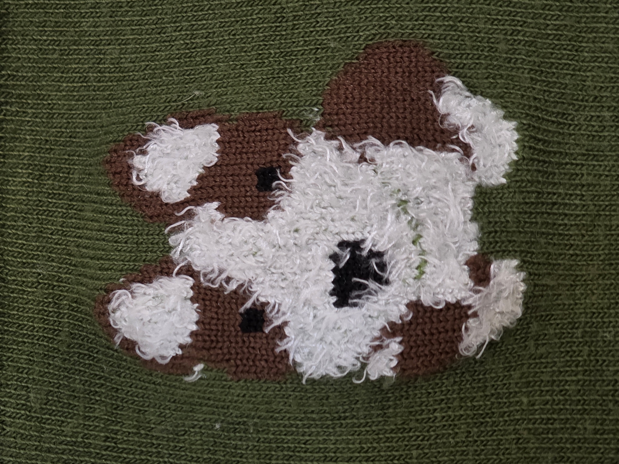 The media file presents a detailed, close-up photograph of a textile object. The primary subject is a multi-colored knitted appliqué, resembling a stylized animal, possibly a dog or a bear cub. This appliqué is set against a background of solid, dark olive-green knitted fabric.
The green background displays a consistent pattern of tight, horizontally oriented stitches, suggesting a fine knit structure like stockinette or a subtle rib. The appliqué itself features three distinct colors and textures. The dominant sections are knitted in a rich brown yarn with clearly defined stitches. Interspersed with these brown areas are significant patches of white yarn, which are notably fluffy and highly textured, likely created using a bouclé or similar yarn type, giving a shaggy or woolly appearance. Within these white and brown sections, two small, square-shaped knitted elements in black yarn are visible, resembling eyes or prominent spots.
No people are visible, and no specific activity or event is taking place; the image is a still life focusing on the craftsmanship and texture of the textile. There is no visible text, and the image itself does not provide cues for time of day, weather, or emotions. The overall impression is of a warm, handmade item, possibly part of a garment like a sweater or a decorative blanket. The provided location context of Kazan, Russia, is not visually discernable from the image.