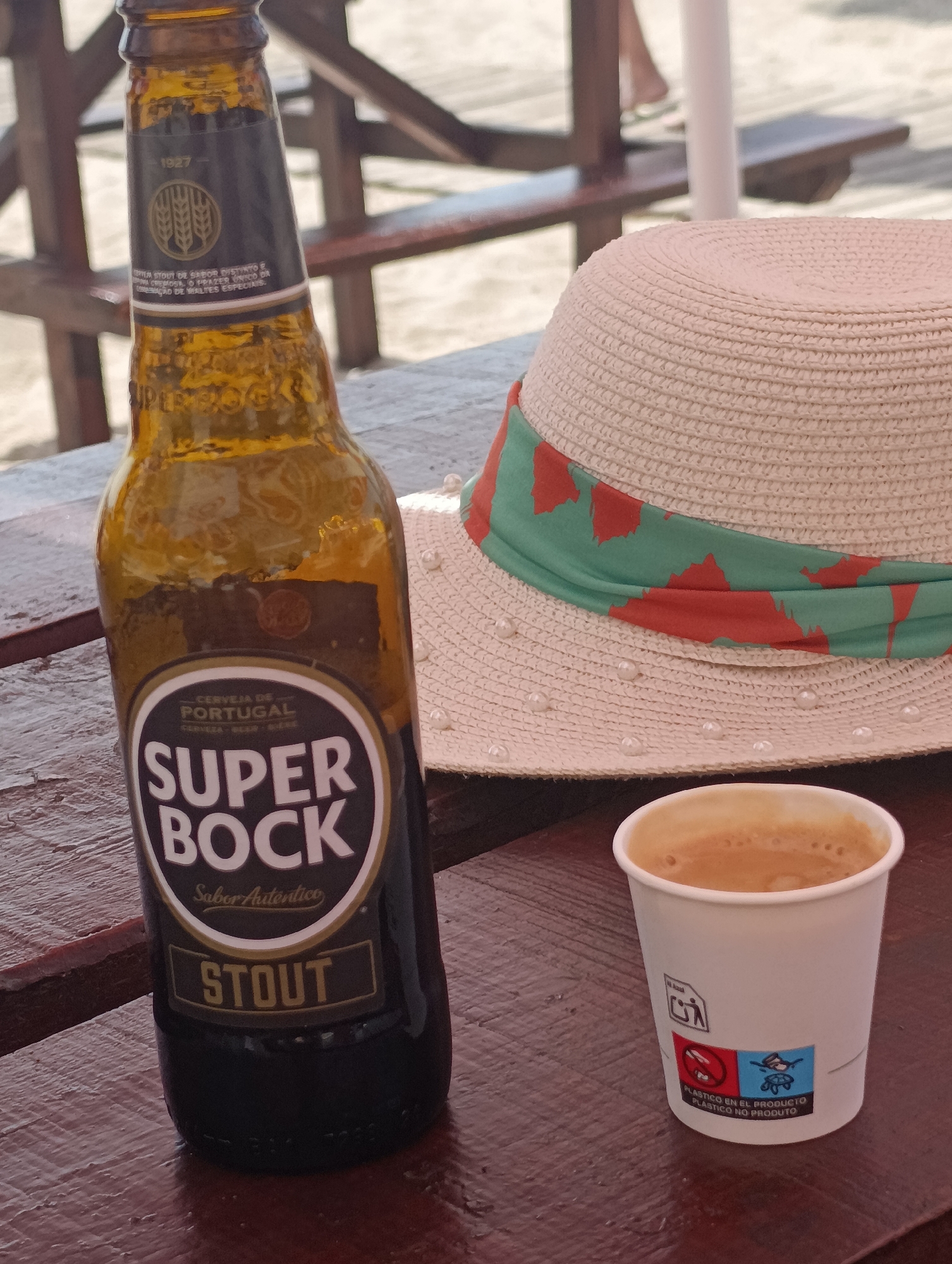A close-up, eye-level shot shows a brown glass bottle of Super Bock Stout beer and a white paper cup of coffee on a dark wooden table. The bottle is on the left, and the cup is on the right. Behind the bottle, a beige straw hat with a green and orange band and decorative pearls sits on the table. In the background, out of focus, are wooden structures, sand, and legs of people, suggesting an outdoor setting like a beach or boardwalk cafe. The lighting is natural and bright, indicating daytime. The image is likely taken at a cafe or bar near the beach in Setúbal, Portugal, given the prominent Portuguese branding on the beer bottle.