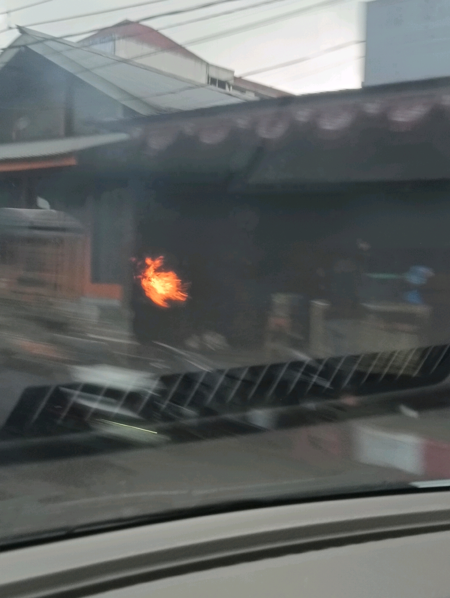 A blurry image taken through a car windshield shows a fire burning in what appears to be an outdoor market or commercial area in Tomohon, Indonesia. The flames are orange and yellow, and they are visible behind a dark, reflective surface. The reflection shows a row of what looks like roof structures with decorative edges. In the foreground, the windshield wipers are visible, indicating the image was taken from inside a moving vehicle. The background is out of focus and shows buildings with sloping roofs and utility lines overhead. The overall impression is one of chaos and destruction due to the fire.